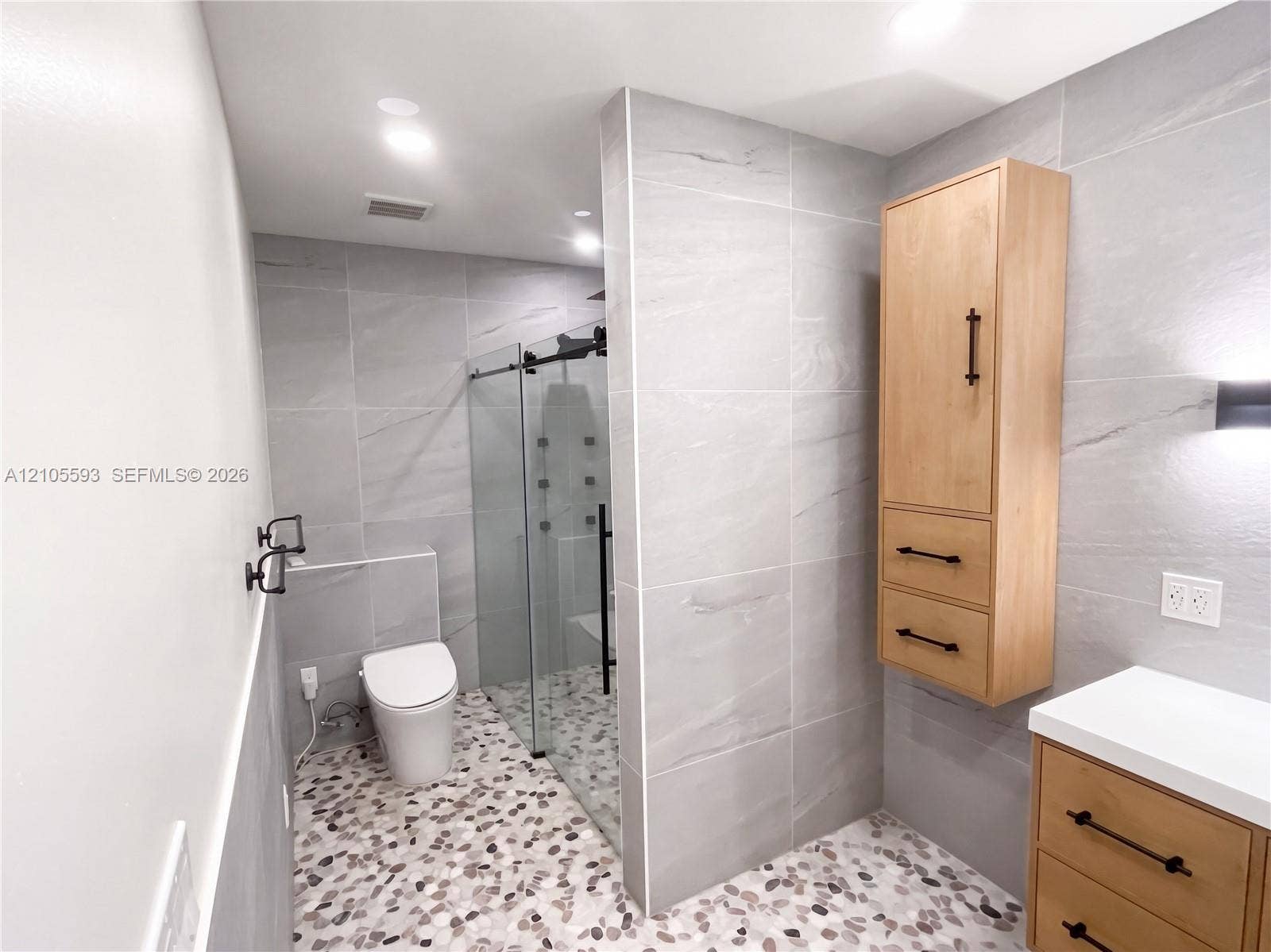 597 Grace Street, Sebastian, FL 32958 Bathroom featuring large-format gray wall tile, a glass-enclosed shower with multiple spray heads, pebble-style flooring, wall-mounted wood-finish cabinetry, and a white countertop