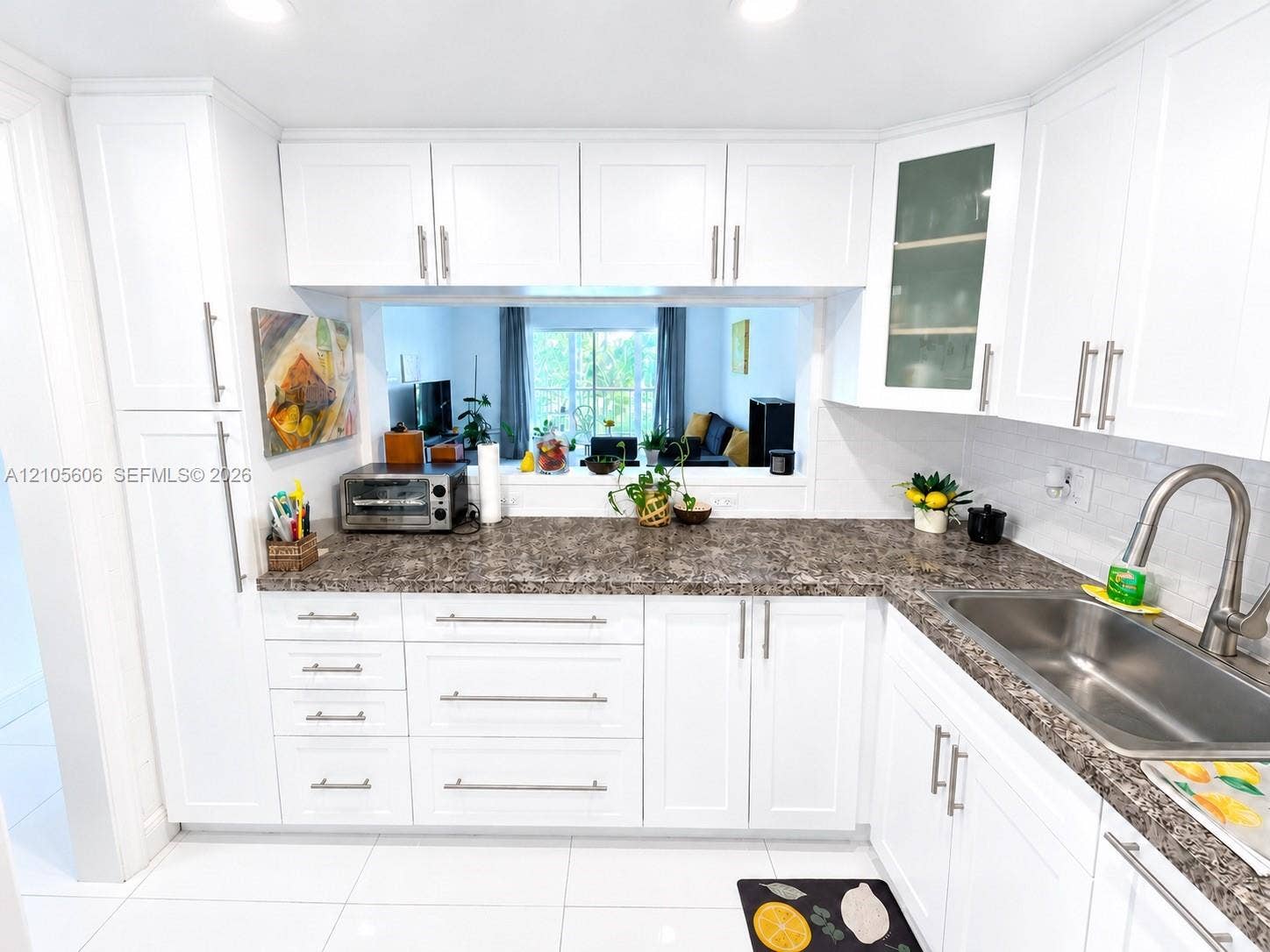 White shaker cabinetry, stone-finish countertops, white subway tile backsplash, stainless steel sink, and a pass-through window