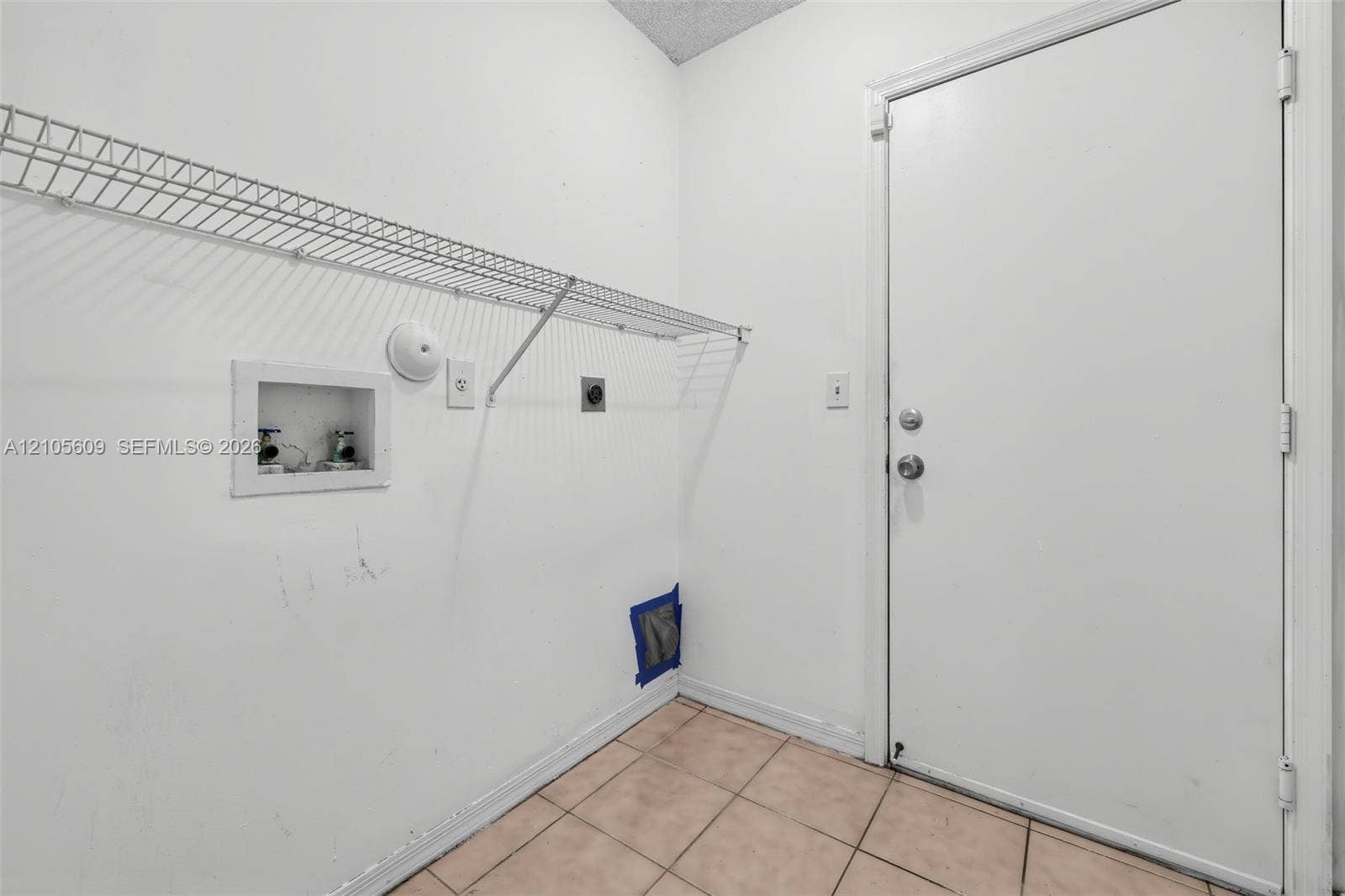 Utility space featuring tiled flooring, a white metal door, and a built-in wire shelving unit with recessed water supply and drainage hookups