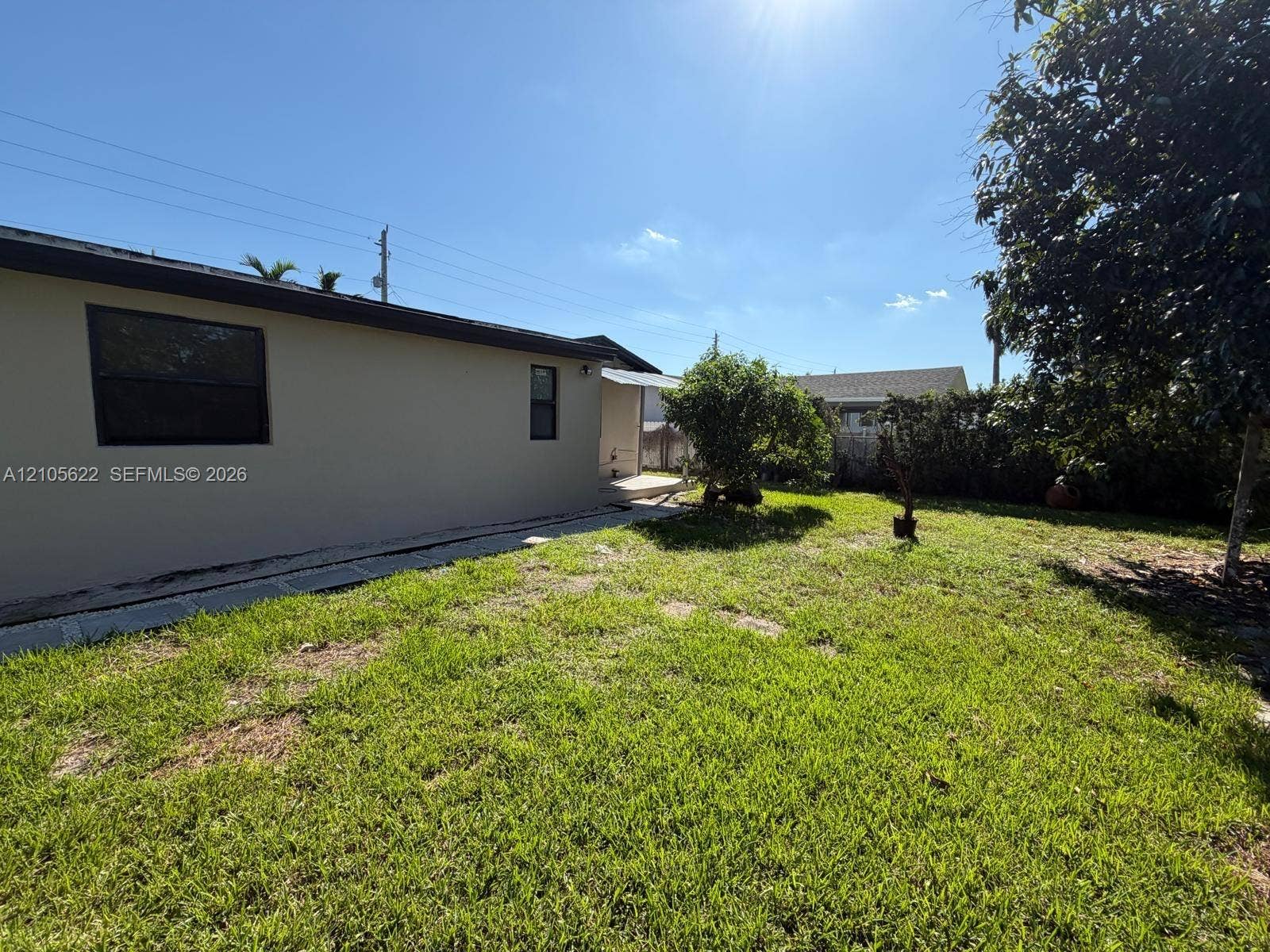 9728 Little River Drive, Miami, FL 33147 Rear exterior featuring a beige stucco facade, dark-trimmed windows, a stone-lined foundation perimeter, and a grass-covered backyard with mature trees