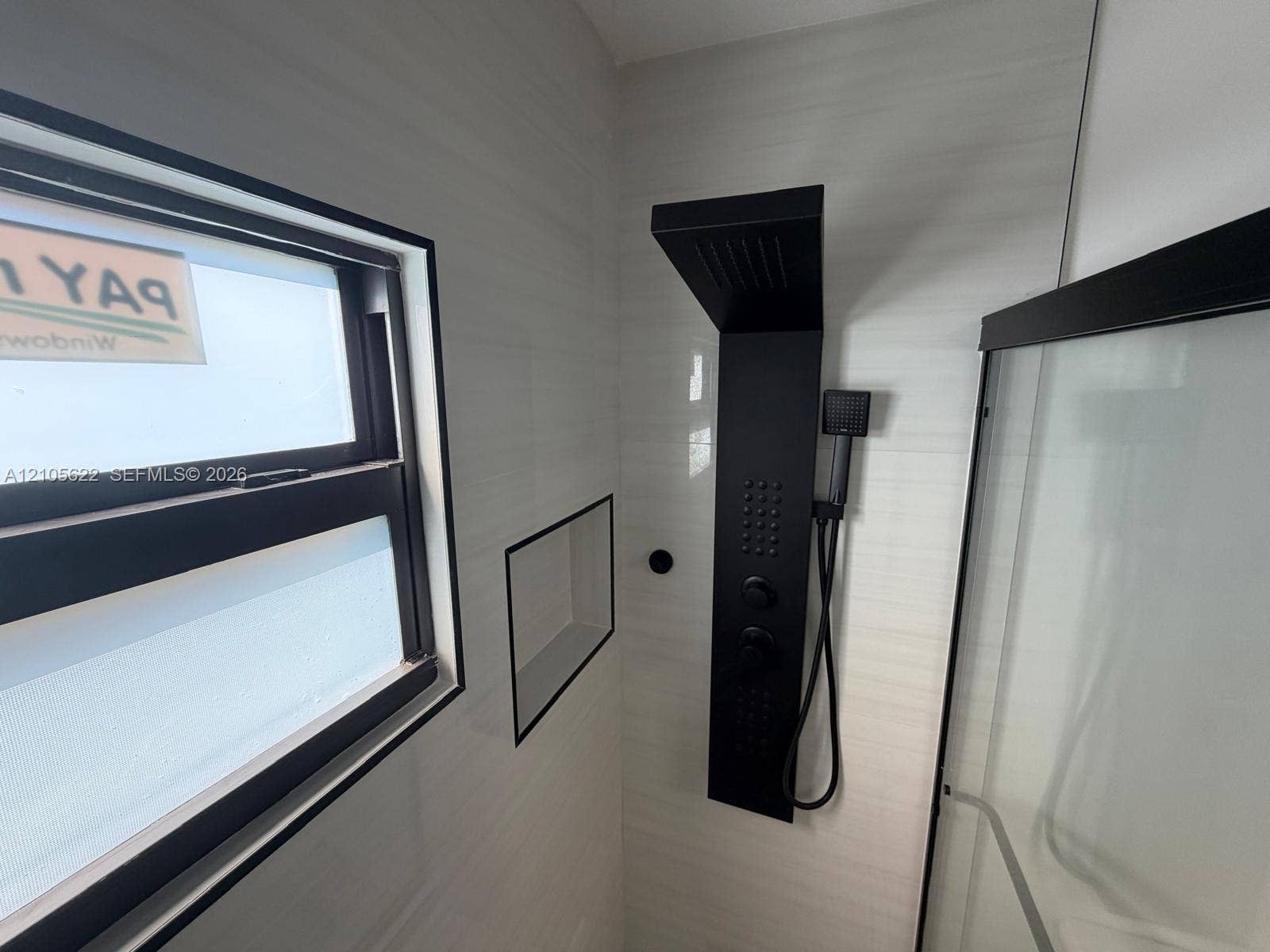 9728 Little River Drive, Miami, FL 33147 Walk-in shower featuring light-toned tile walls, a matte black multi-function shower panel with an integrated handheld sprayer, a built-in recessed wall niche, and a window with a matte black frame