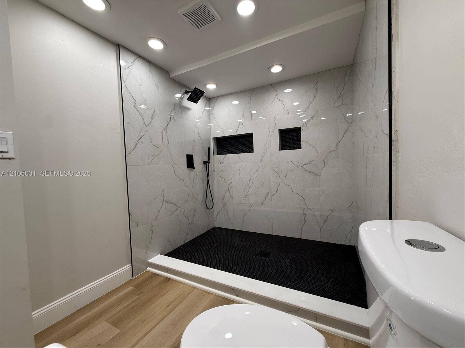 610 NW 13th Street, #0140, Boca Raton, FL 33486 Walk-in shower featuring large-format veined tile surround, matte black rainfall showerhead, integrated wall niches, and black mosaic tile flooring, finished with wood-finish flooring