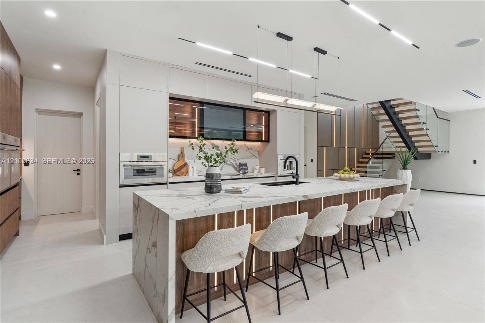 623 NE 16th Terrace, Fort Lauderdale, FL 33304 Modern kitchen featuring a waterfall-edge island with stone-finish surfaces and integrated wood-paneling, flat-panel cabinetry, built-in stainless steel appliances, and a staircase with a glass railing and wood treads