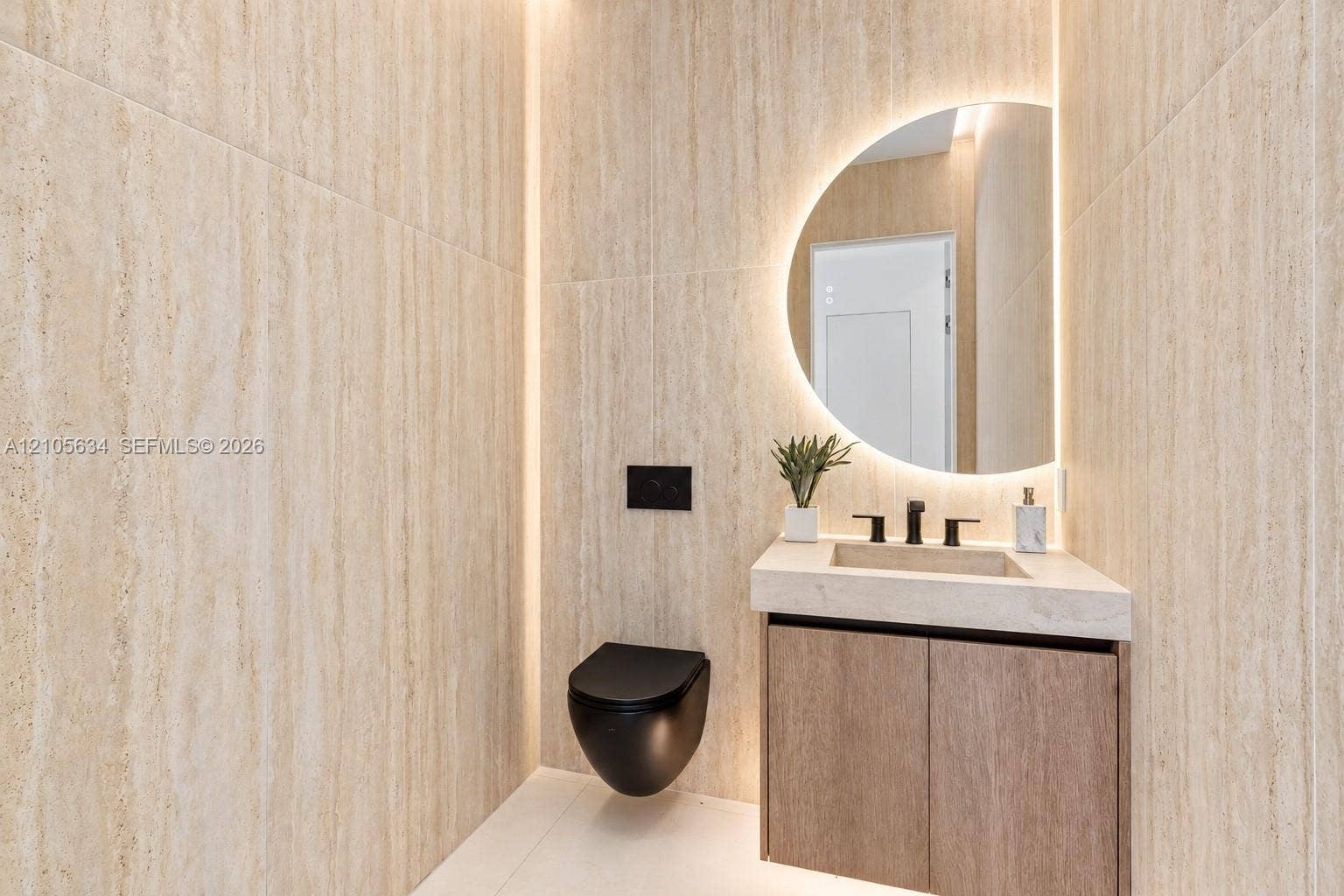 623 NE 16th Terrace, Fort Lauderdale, FL 33304 Full-height stone-finish wall tiling, wall-mounted matte black toilet, floating wood-finish vanity with integrated stone-finish basin, matte black widespread faucet, and semi-circular backlit wall mirror
