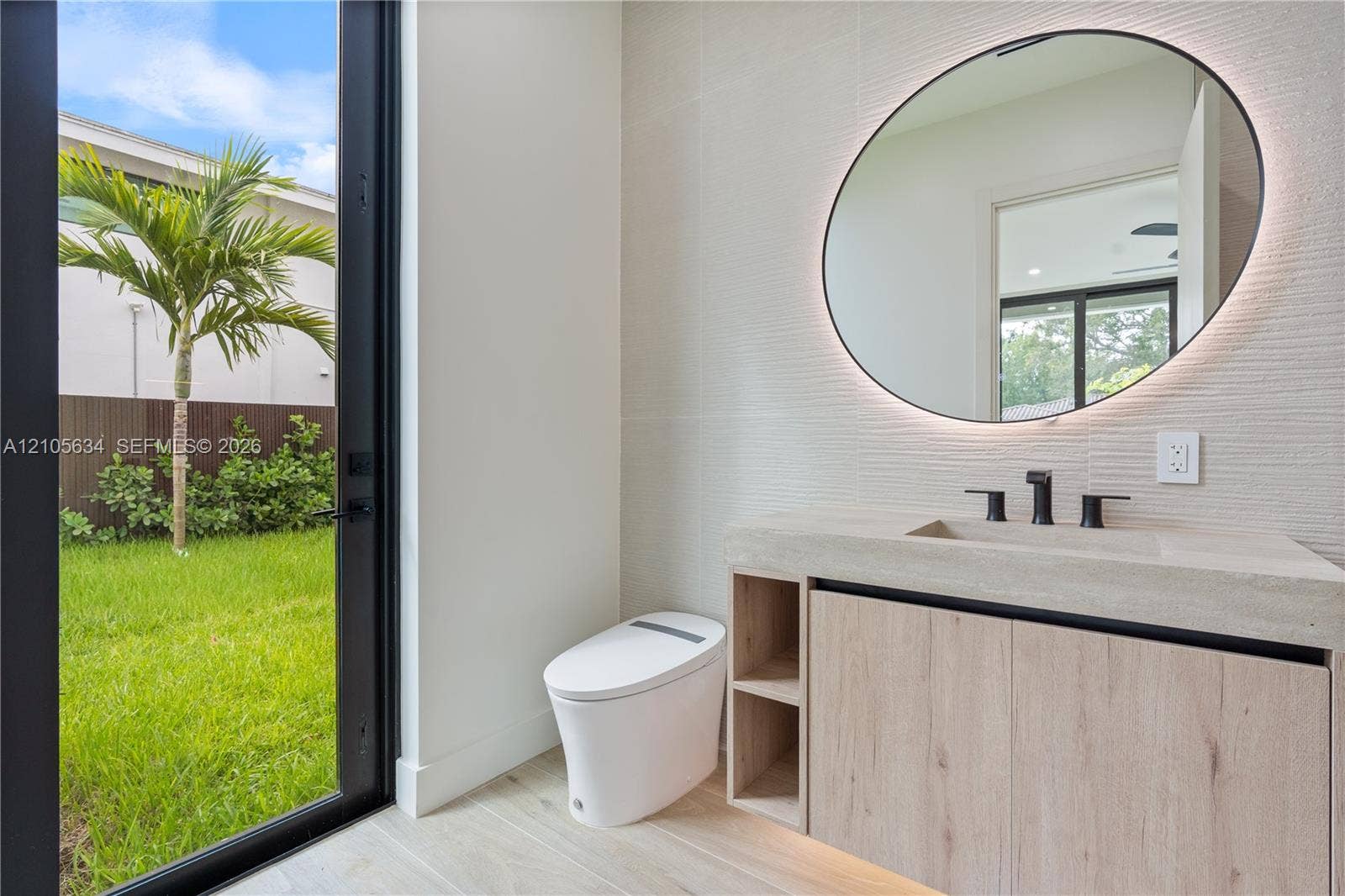 623 NE 16th Terrace, Fort Lauderdale, FL 33304 Modern vanity with stone-finish countertop, integrated sink, and wood-finish cabinetry, featuring a circular backlit mirror, wall-mounted matte black faucet, and full-height glass door