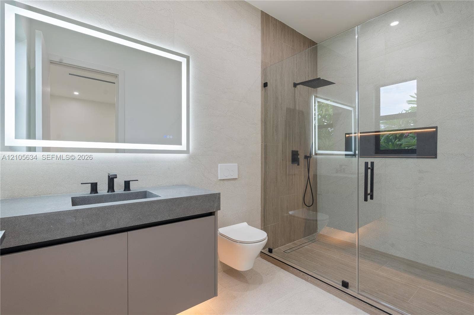 623 NE 16th Terrace, Fort Lauderdale, FL 33304 Floating vanity with integrated sink and matte black faucet, wall-mounted toilet, walk-in shower with glass enclosure, wood-finish tile shower wall, and illuminated vanity mirror
