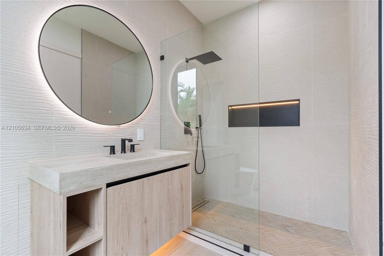 623 NE 16th Terrace, Fort Lauderdale, FL 33304 Floating vanity with integrated sink, textured wall tile, circular backlit mirror, frameless glass shower enclosure with rainfall showerhead, and herringbone-patterned shower floor tile