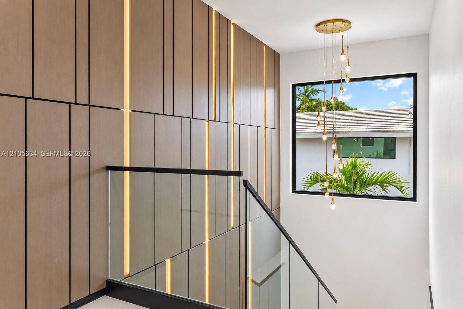 623 NE 16th Terrace, Fort Lauderdale, FL 33304 Custom wood-panelled accent wall with integrated vertical lighting, glass railing system featuring a black metal handrail, and multi-pendant brass chandelier