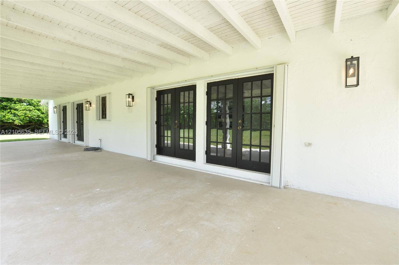 16760 SW 280th Street, Homestead, FL 33031 Covered patio featuring concrete flooring, white textured wall finish, exposed beam ceiling, two sets of multi-pane French doors with dark frames, and wall-mounted exterior lanterns