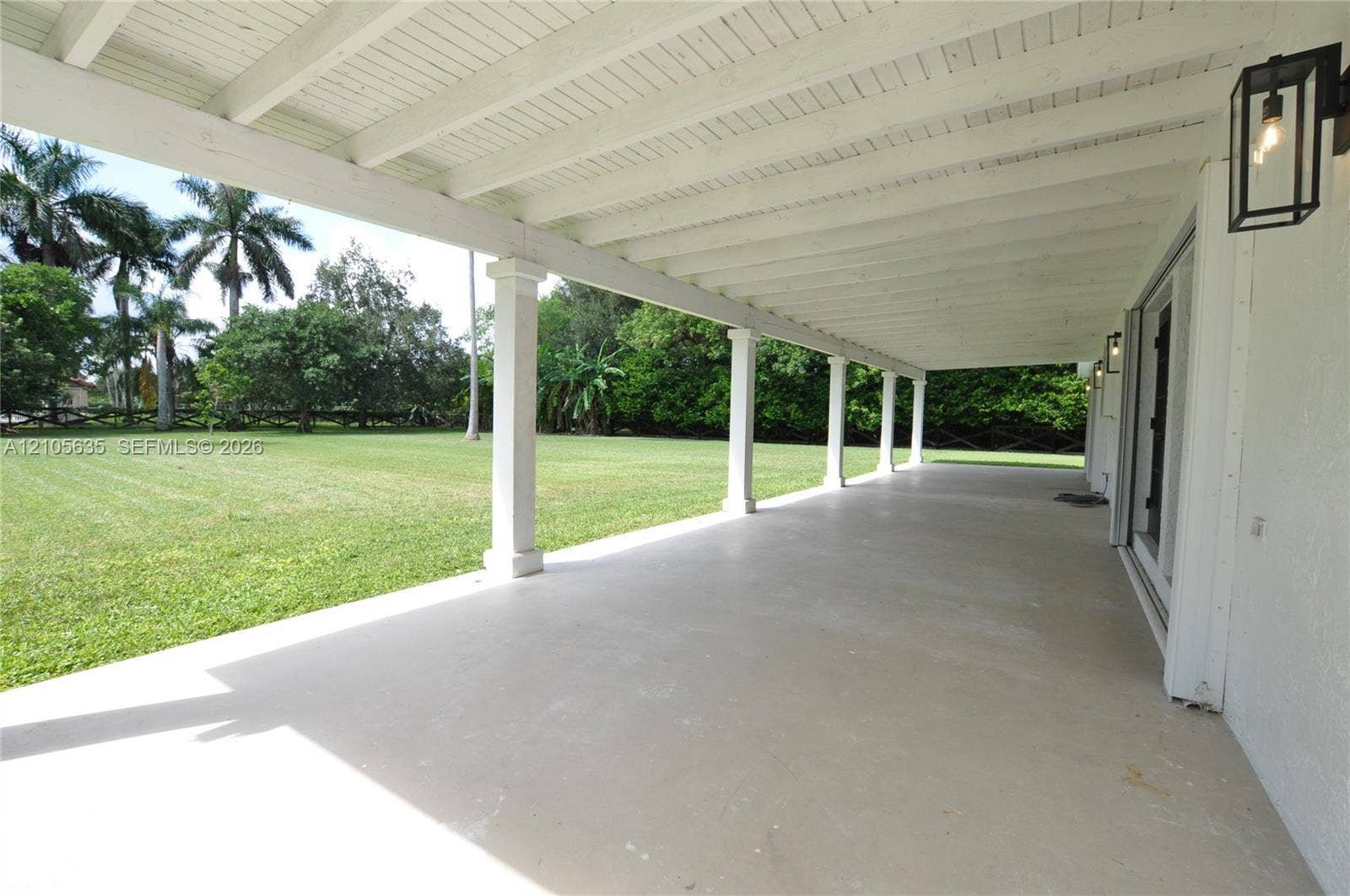 16760 SW 280th Street, Homestead, FL 33031 Covered patio featuring a painted wood-beam ceiling, structural columns, sleek concrete flooring, and minimalist wall-mounted light fixtures, overlooking an expansive manicured lawn