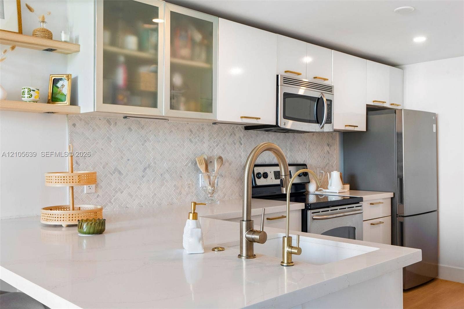 Kitchen featuring white cabinetry with glass-front uppers, herringbone-patterned stone backsplash, quartz countertops, brass-finish fixtures, and built-in stainless steel appliances