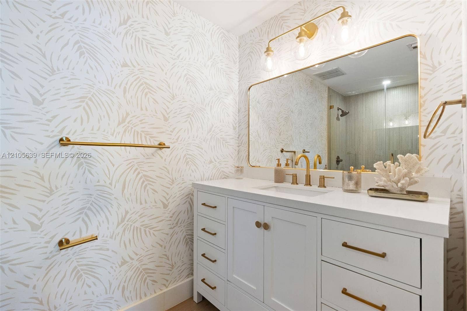 Shaker-style white vanity with gold-tone hardware, rectangular undermount sink, solid-surface countertop, gold-tone vanity lighting, and rectangular mirror with a gold-tone frame