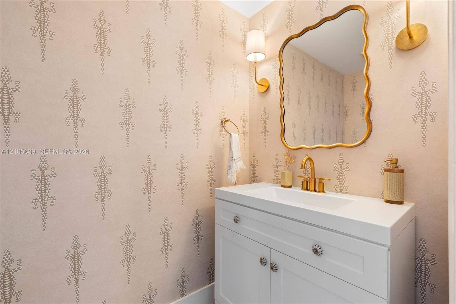 Patterned wall covering, white vanity cabinet with decorative hardware, white rectangular sink, gold-toned faucet and hardware, and wavy-edged mirror with gold-toned frame