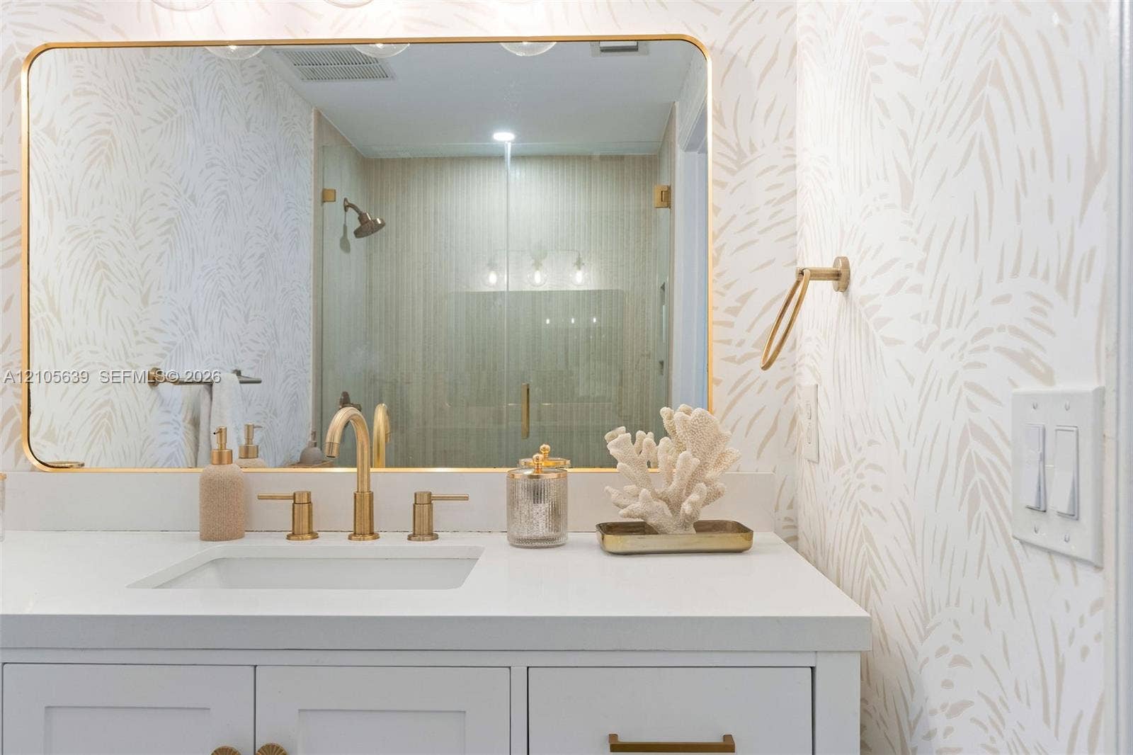 Vanity featuring white shaker-style cabinetry, a solid surface countertop, brass-toned hardware, an integrated rectangular basin, and a gold-framed wall mirror