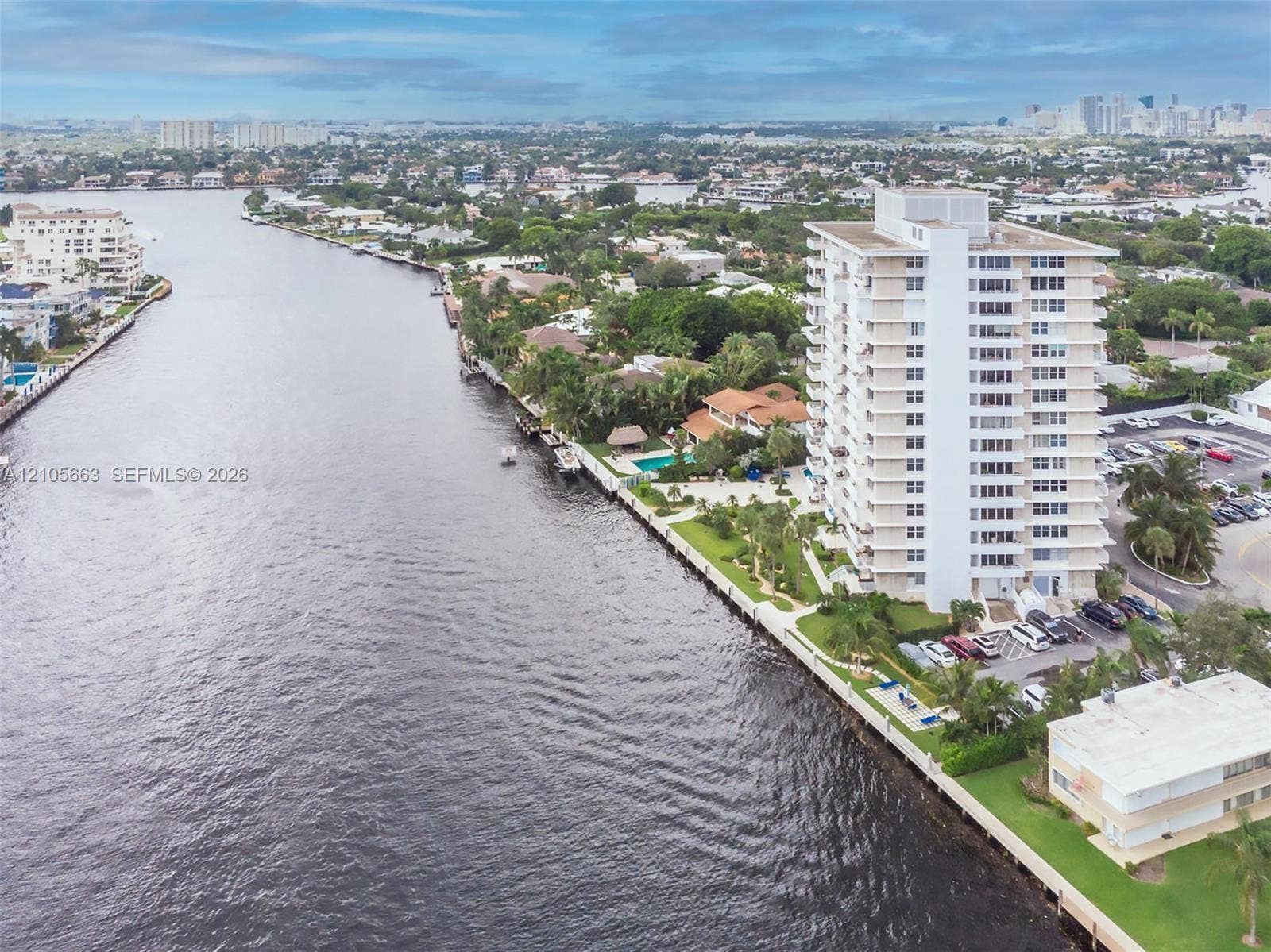 Aerial perspective of high-rise building situated on a waterway with bulkhead frontage, landscaped grounds, surface parking, and urban skyline backdrop