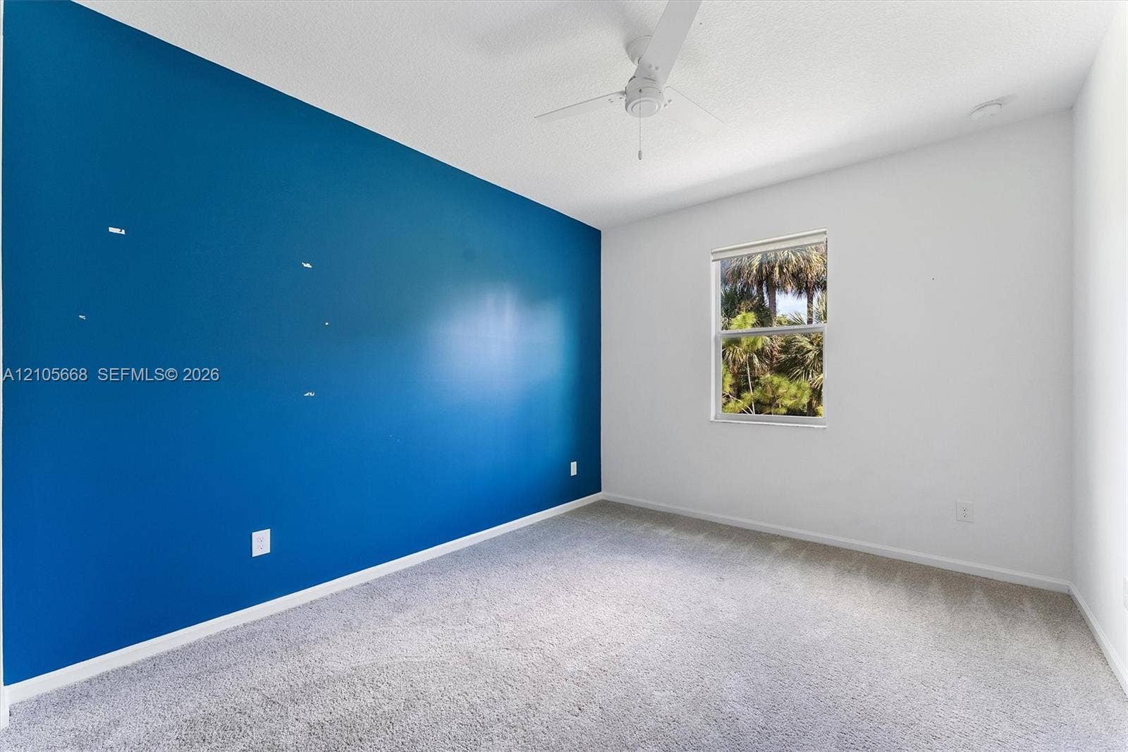 6307 Trails Of Foxford Court, West Palm Beach, FL 33415 Interior space featuring a vibrant blue accent wall, neutral carpeting, a multi-blade ceiling fan, and a single window with exterior greenery