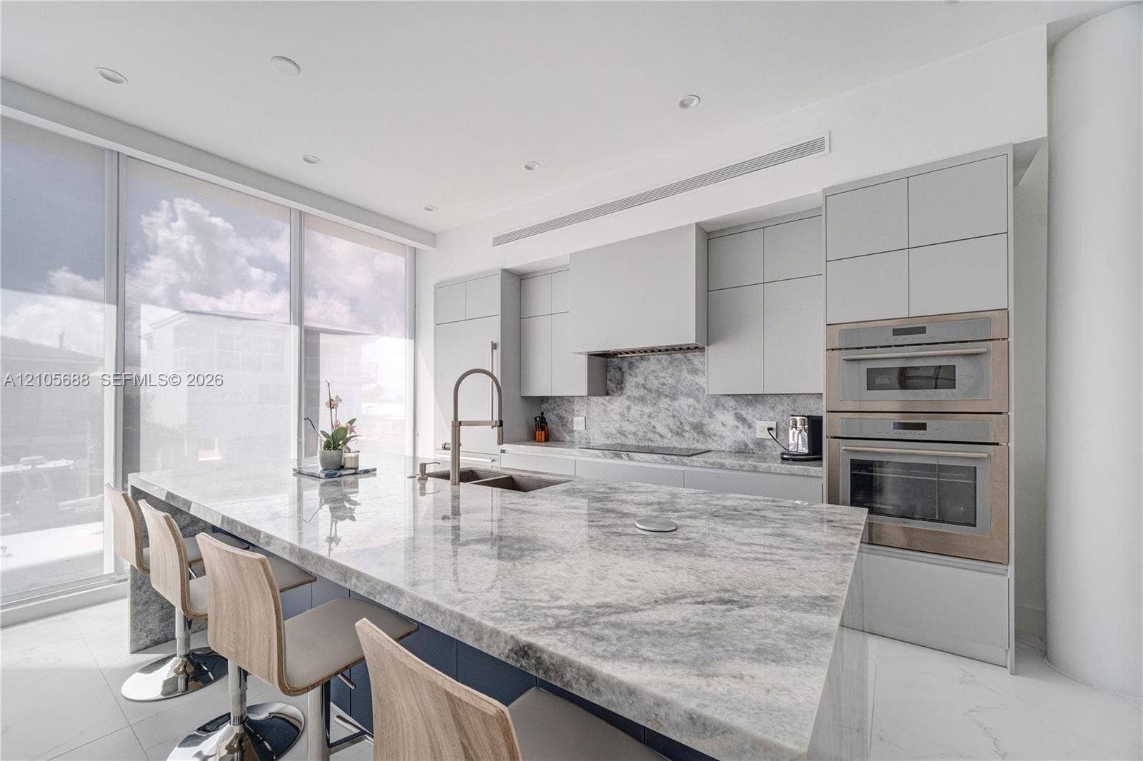 164 W Avenue C, Key Largo, FL 33037 Modern kitchen featuring floor-to-ceiling windows, a large stone-surfaced waterfall island, matching stone slab backsplash, flat-panel cabinetry, and integrated stainless steel double wall ovens
