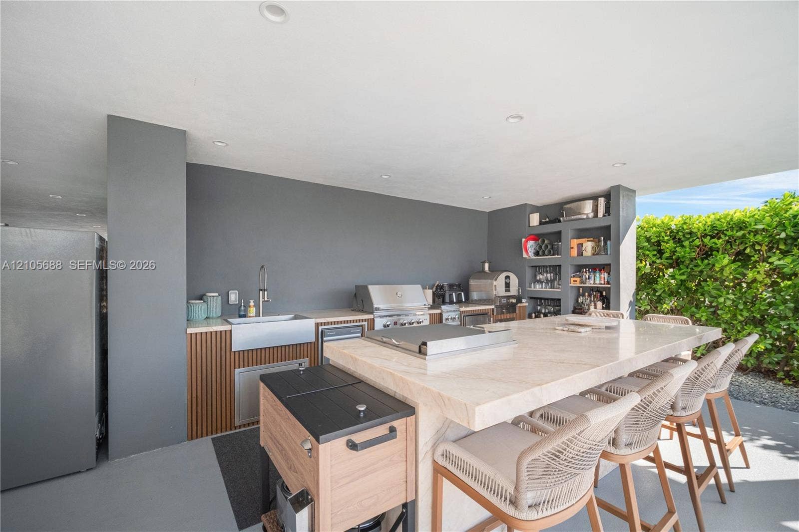 164 W Avenue C, Key Largo, FL 33037 Covered outdoor kitchen featuring a stone-topped island, stainless steel farmhouse sink, built-in stainless steel grill, and custom wood-slat cabinetry with integrated shelving