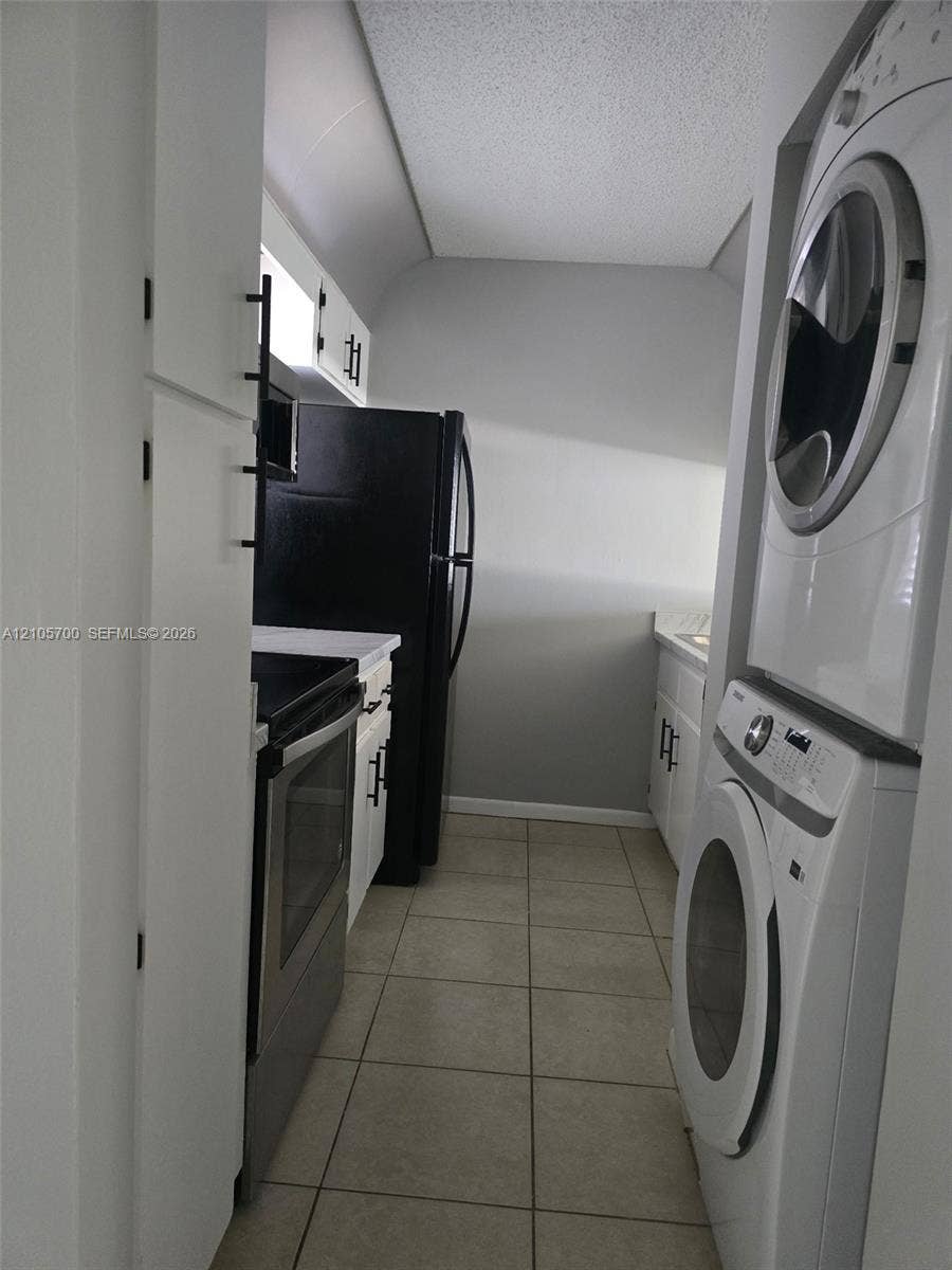 Utility space featuring large-format tile flooring, stacked front-load laundry appliances, white cabinetry with matte black hardware, and an integrated refrigerator and range