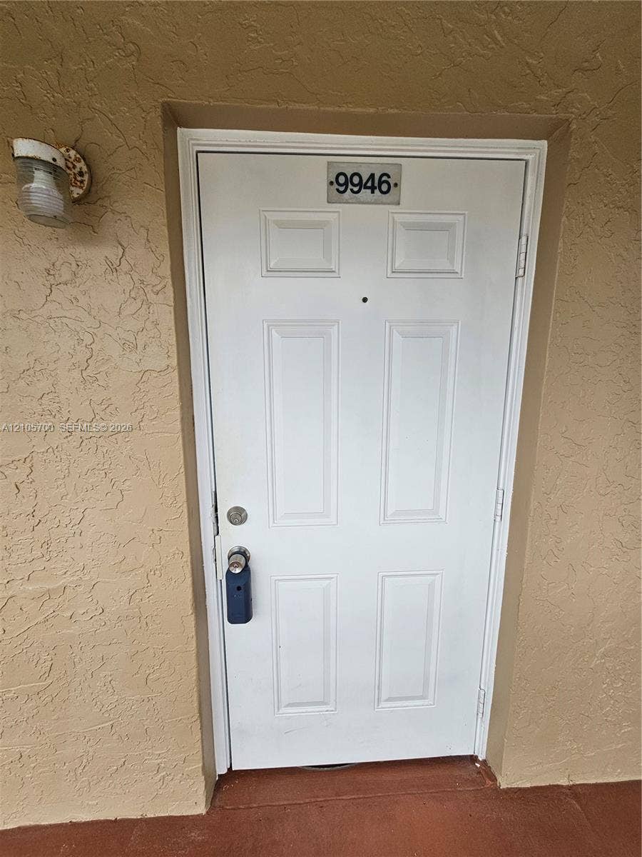 White six-panel entry door featuring a center-mounted peephole, metal address plaque, and wall-mounted exterior light fixture