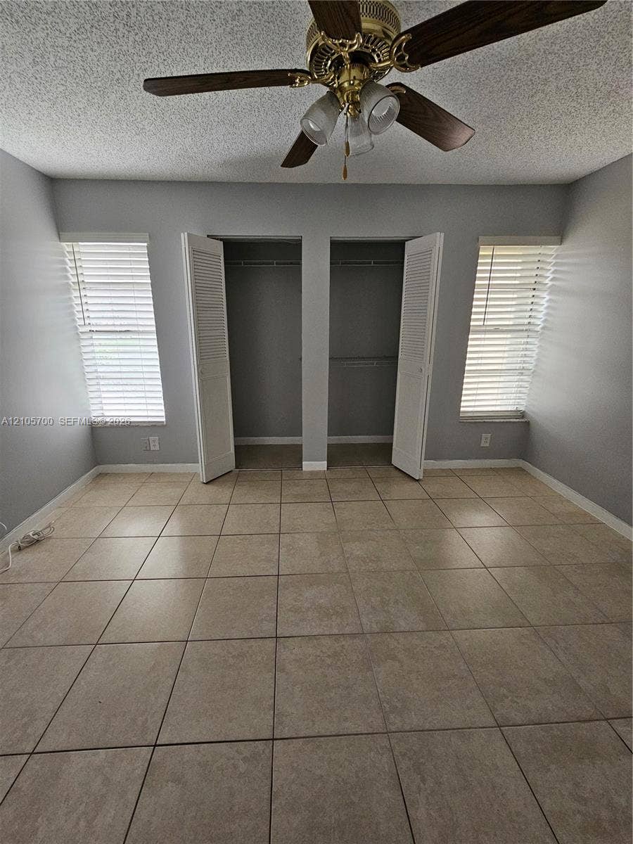 Room featuring neutral-toned floor tiling, two separate closets with louvered doors, twin windows with horizontal blinds, and an overhead ceiling fan