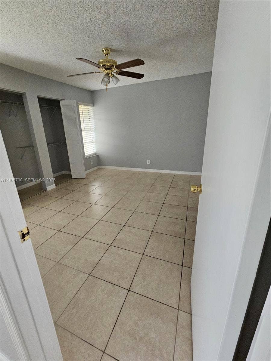 Room interior featuring large-format ceramic tile flooring, neutral wall paint, dual closets with louvered doors, and a ceiling fan with integrated lighting