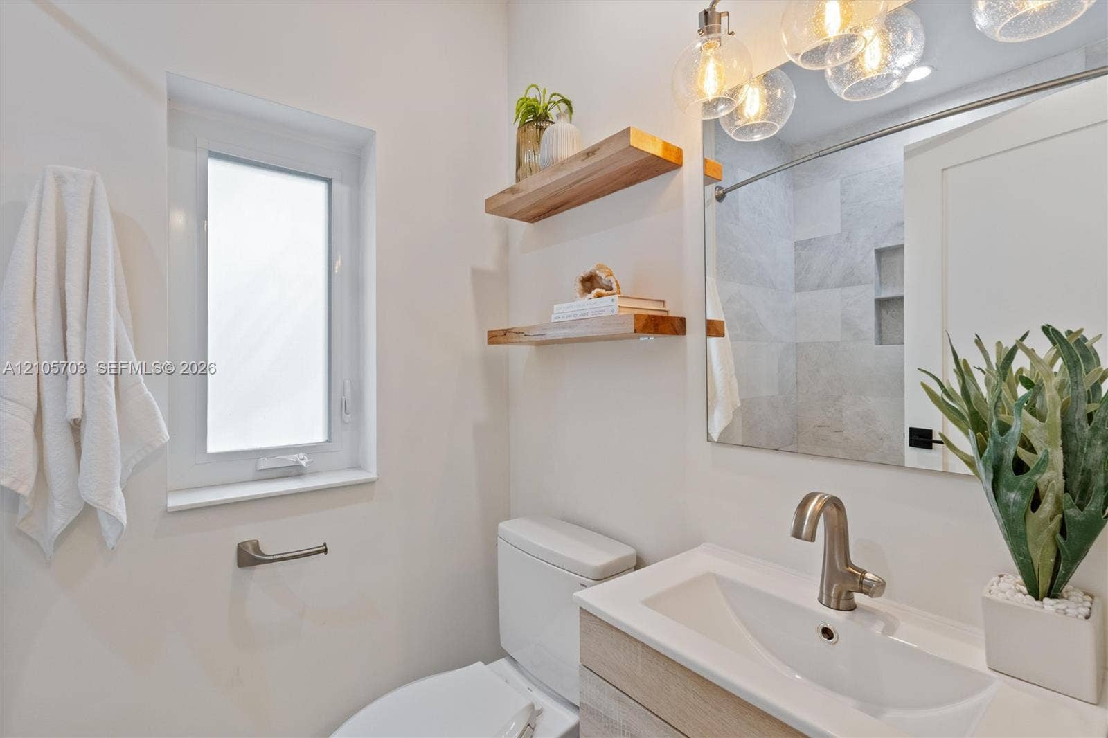 190 NW 102nd Street, Miami Shores, FL 33150 Bathroom featuring a light-toned wood-finish vanity with a rectangular sink, wall-mounted natural wood shelving, multi-bulb globe pendant lighting, and a walk-in shower with tiled surround