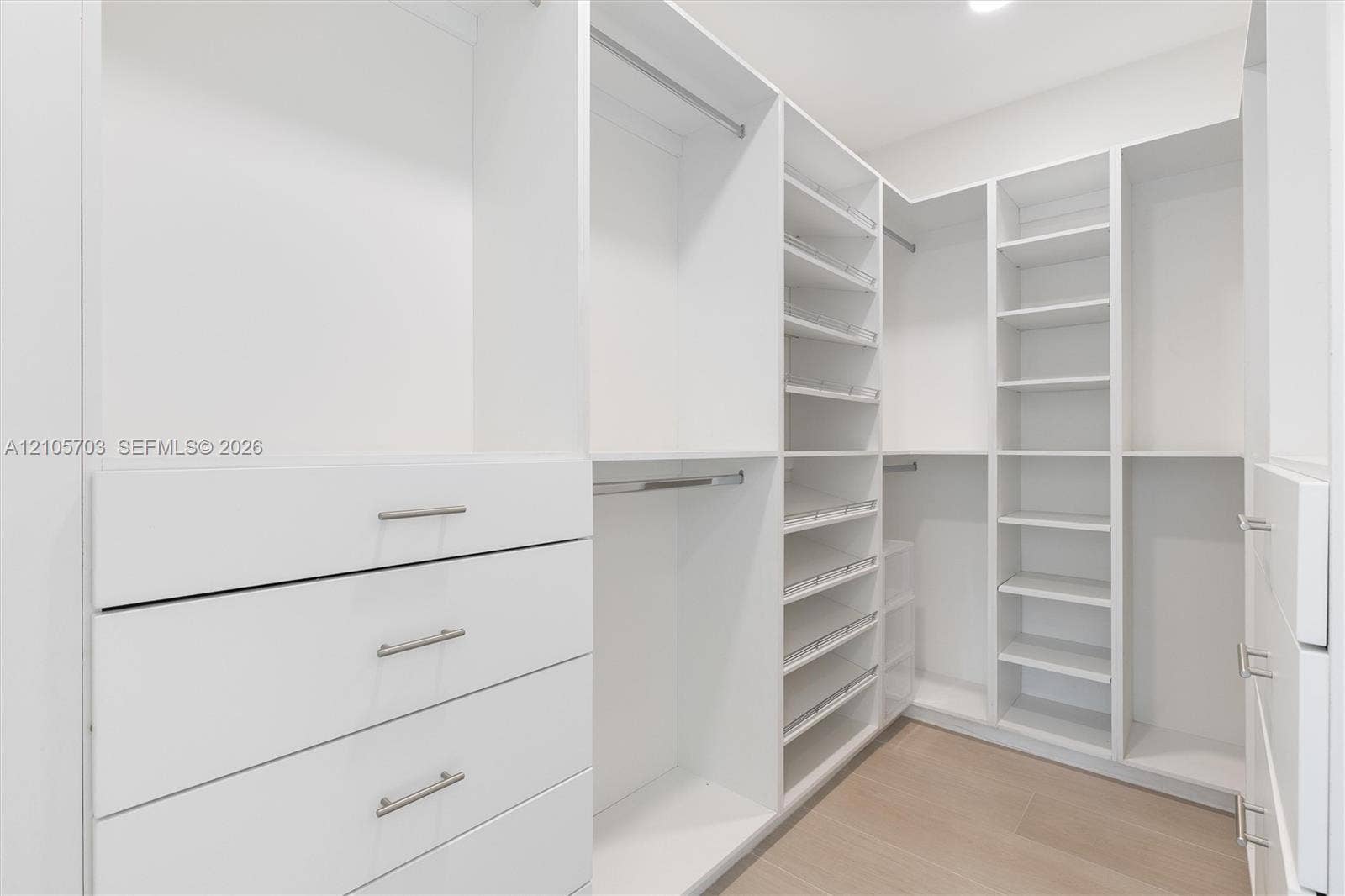 190 NW 102nd Street, Miami Shores, FL 33150 Luxurious walk-in closet featuring custom white cabinetry with integrated drawers, extensive shelving throughout, polished chrome hardware, and warm wood-finish flooring
