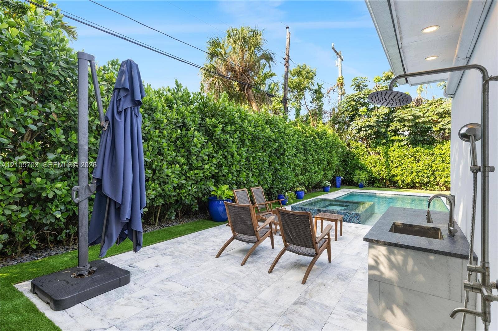 190 NW 102nd Street, Miami Shores, FL 33150 Outdoor patio featuring natural stone paving, an integrated wet bar with a dark stone countertop, a wall-mounted shower fixture, an in-ground swimming pool with an elevated spa, and mature privacy hedging