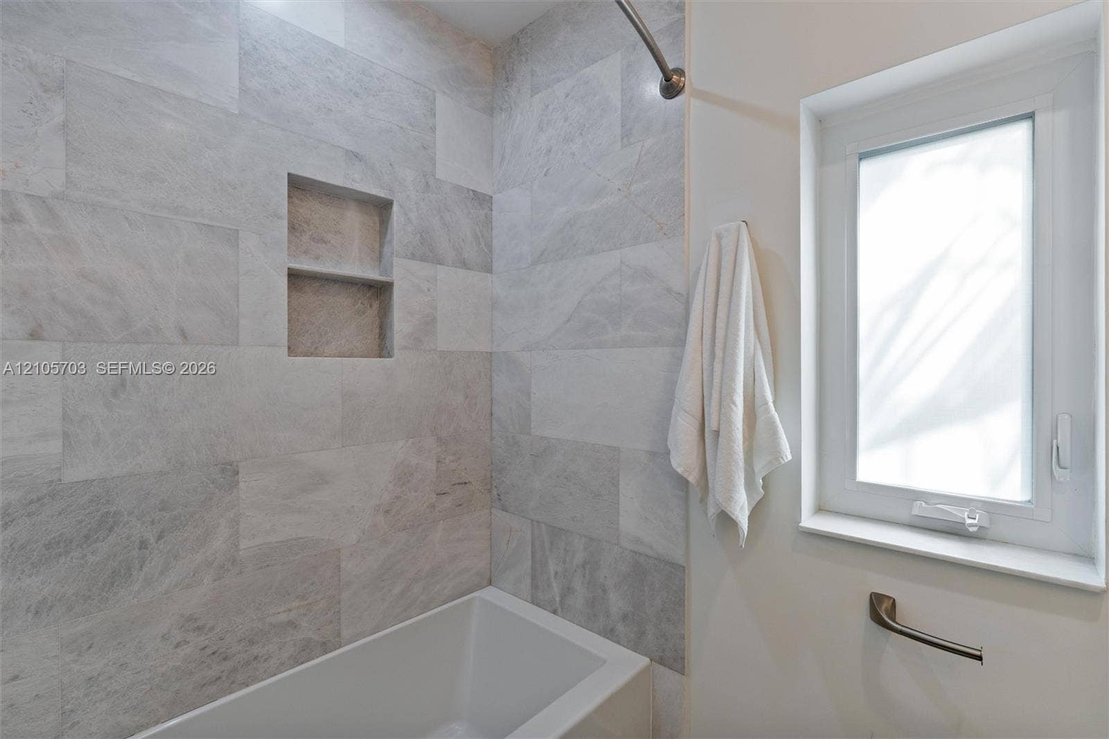 190 NW 102nd Street, Miami Shores, FL 33150 Full-height shower surround featuring large-format light grey stone tiling, recessed double-niche shelving, a built-in soaking tub, and a window with a mounted metal grab bar