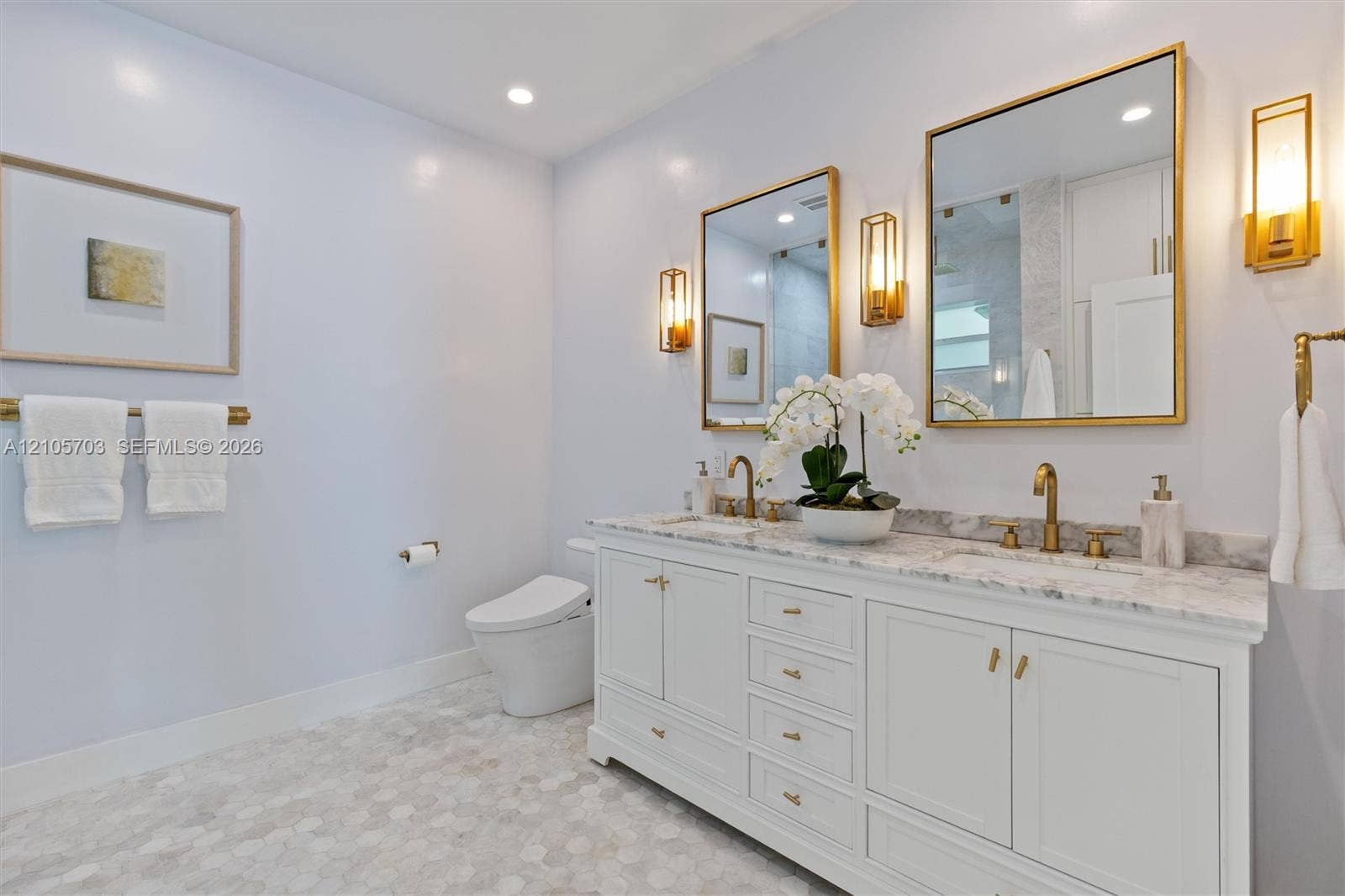 190 NW 102nd Street, Miami Shores, FL 33150 Dual-vanity cabinetry with stone-finish countertops, undermount sinks, brass-toned fixtures, metallic-framed wall mirrors, and wall-mounted sconce lighting