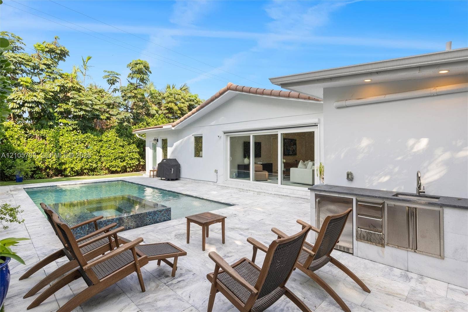 190 NW 102nd Street, Miami Shores, FL 33150 Outdoor living area featuring a custom swimming pool with integrated spa, light-toned stone patio, built-in stainless steel outdoor kitchen with a sink, and recessed exterior lighting