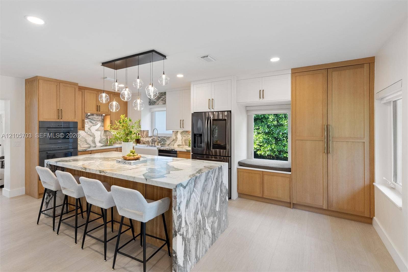 190 NW 102nd Street, Miami Shores, FL 33150 Kitchen featuring a large waterfall island with patterned stone surfacing, natural wood-finish cabinetry, built-in dual ovens, multi-light pendant fixture, and a window-side built-in bench