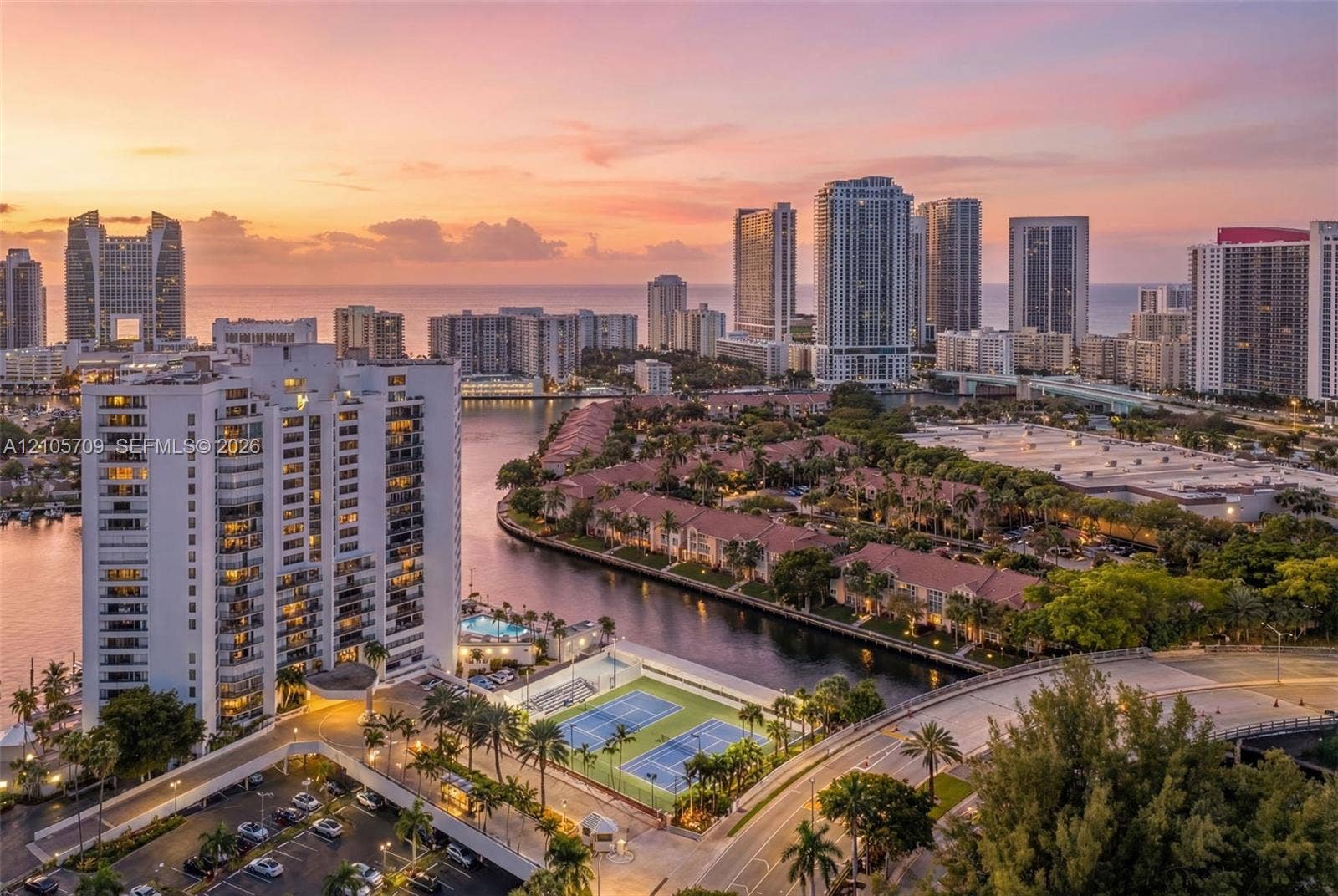 300 Three Islands Boulevard, #516, Hallandale Beach, FL 33009 High-rise residential property featuring balcony access, waterfront placement, and proximity to hard-surface tennis courts and lush tropical landscaping