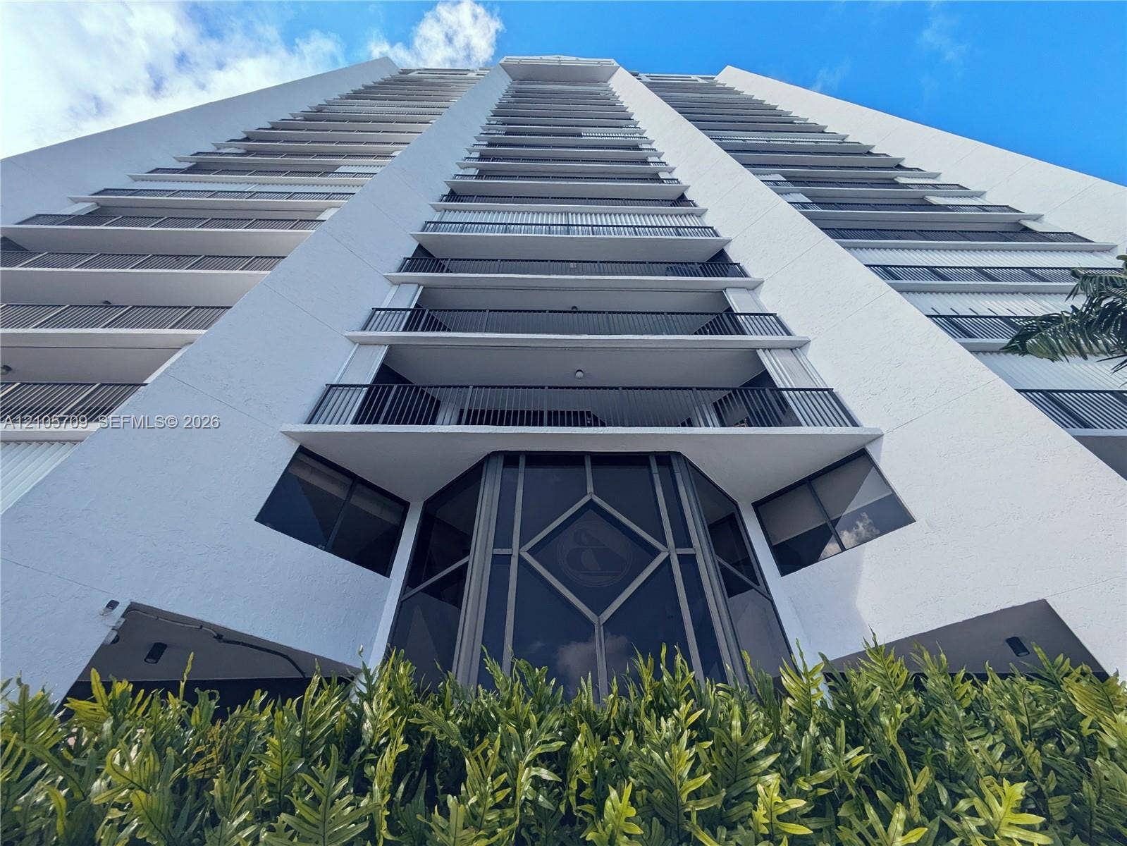 300 Three Islands Boulevard, #516, Hallandale Beach, FL 33009 High-rise building exterior featuring a white concrete faade, tiered private balconies with metal railings, floor-to-ceiling glass paneling, and a geometric-patterned entryway