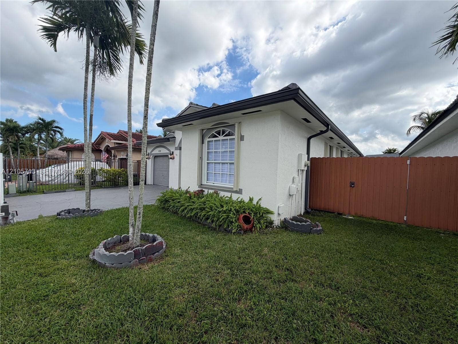 Stucco exterior featuring an arched window, a solid-color wood fence, palm trees, and landscaped shrubbery