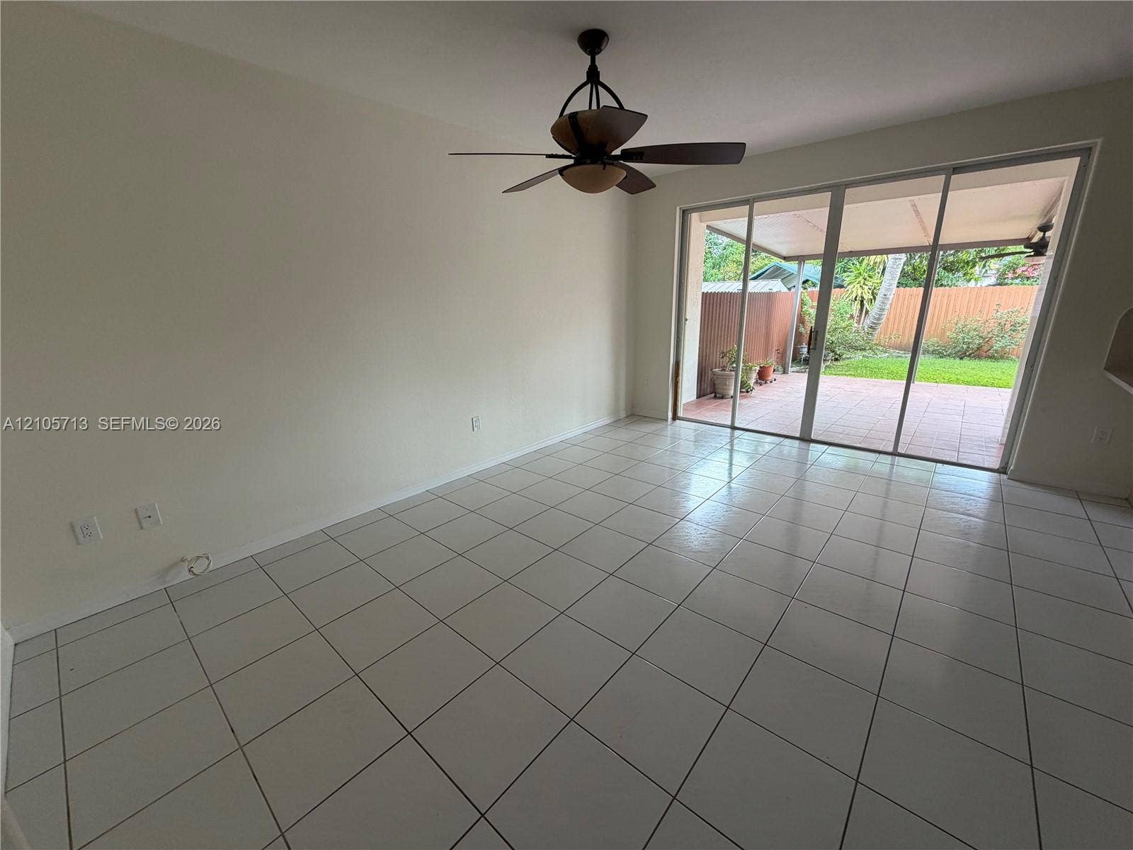 Interior space featuring neutral wall paint, white tile flooring, a ceiling fan with integrated lighting, and full-height sliding glass doors opening to a covered patio and enclosed lawn