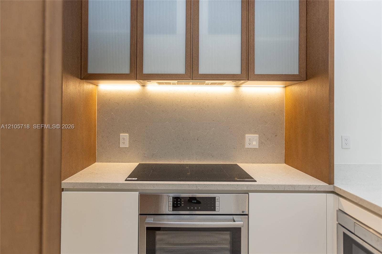2200 Brickell Avenue, #GV16, Miami, FL 33129 Kitchen area featuring matte white base cabinetry, integrated stainless steel oven, cooktop, solid-surface countertop, and wood-finish upper cabinets with fluted glass panel inserts