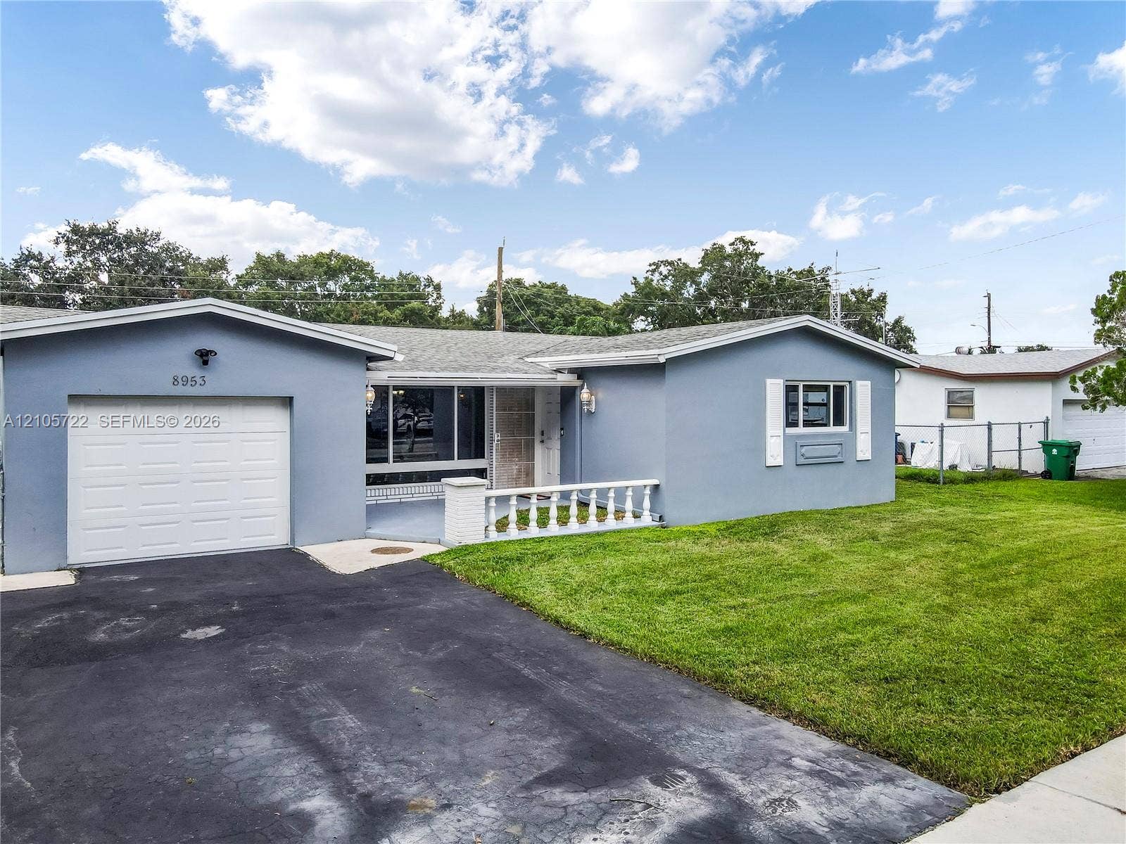 8953 SW 53rd Street, Cooper City, FL 33328 Single-story residence featuring a gray stucco exterior, white louvered shutters, an attached one-car garage, and a decorative white porch railing