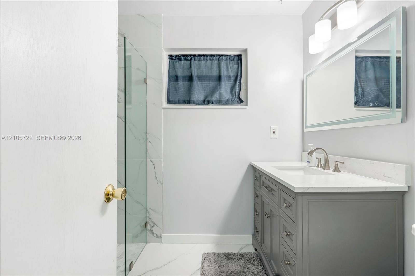 8953 SW 53rd Street, Cooper City, FL 33328 Gray shaker-style vanity featuring a white stone-composite countertop, under-mount sink, chrome faucet, wall-to-wall mirror with integrated lighting, and a glass-enclosed shower with large-format tile surround