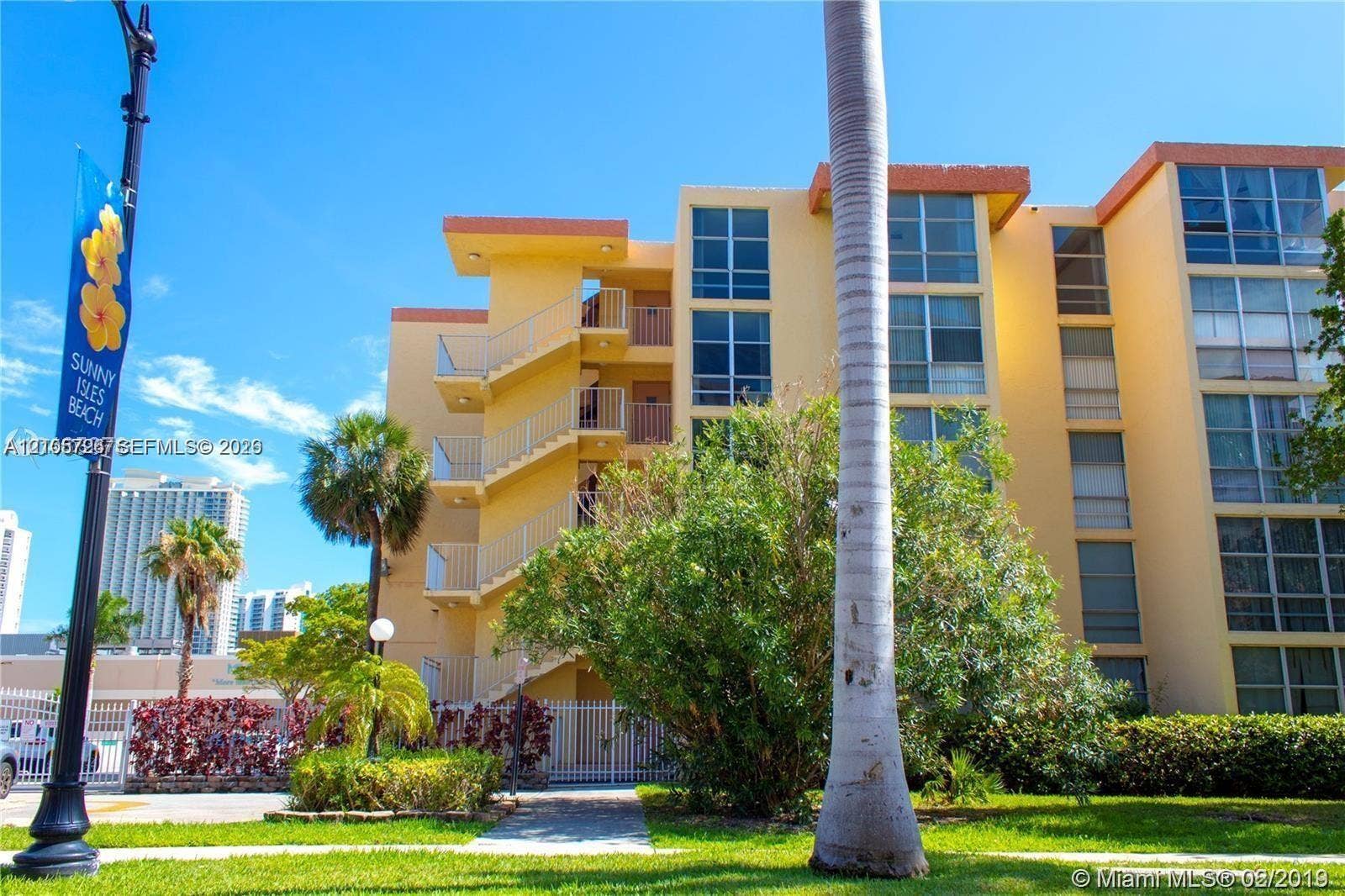 200 172nd Street, #101, Sunny Isles Beach, FL 33160 Multi-story residential building exterior featuring a stucco facade, expansive glass windows, external staircases with metal railings, and landscaped grounds with mature palm trees