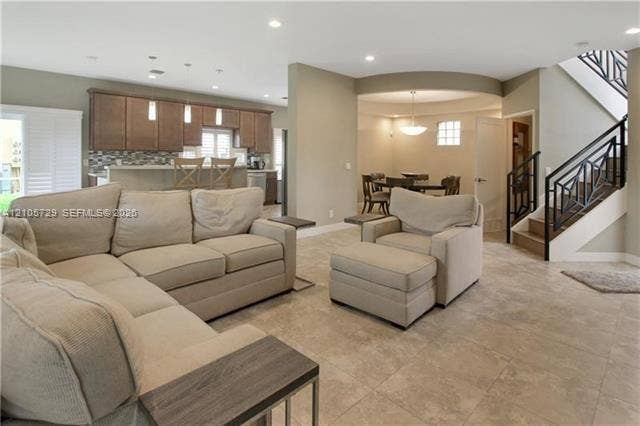 Open-concept interior featuring neutral stone-finish tile flooring, recessed lighting, wood cabinetry, stone-tiled backsplash, and a staircase with decorative metal railing