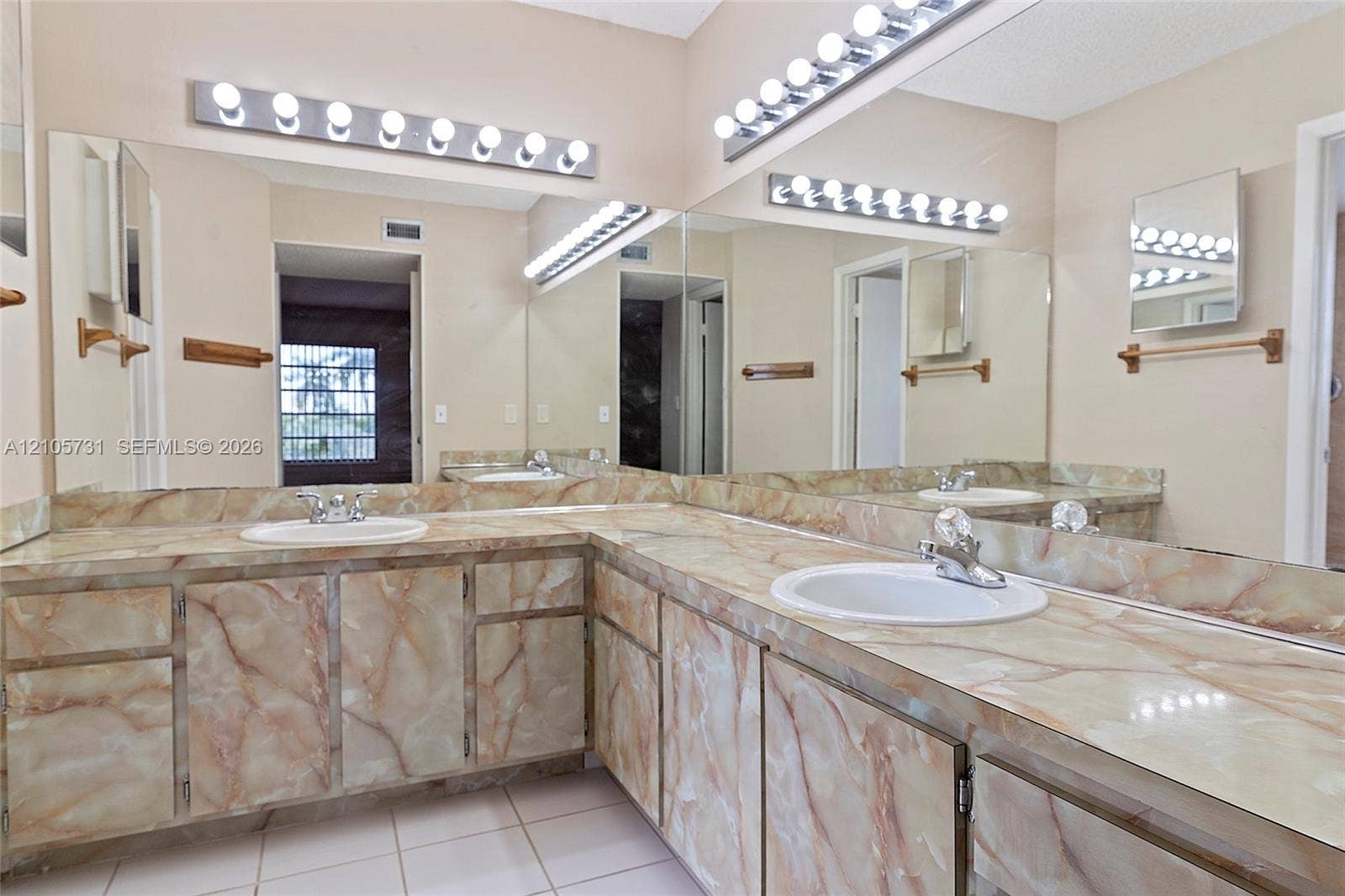 5700 SW 127th Avenue, #1404, Miami, FL 33183 Dual-sink vanity featuring patterned stone-finish countertops, custom cabinetry with matching stone-finish doors, expansive wall-to-wall mirror, and multi-bulb theatrical lighting fixtures