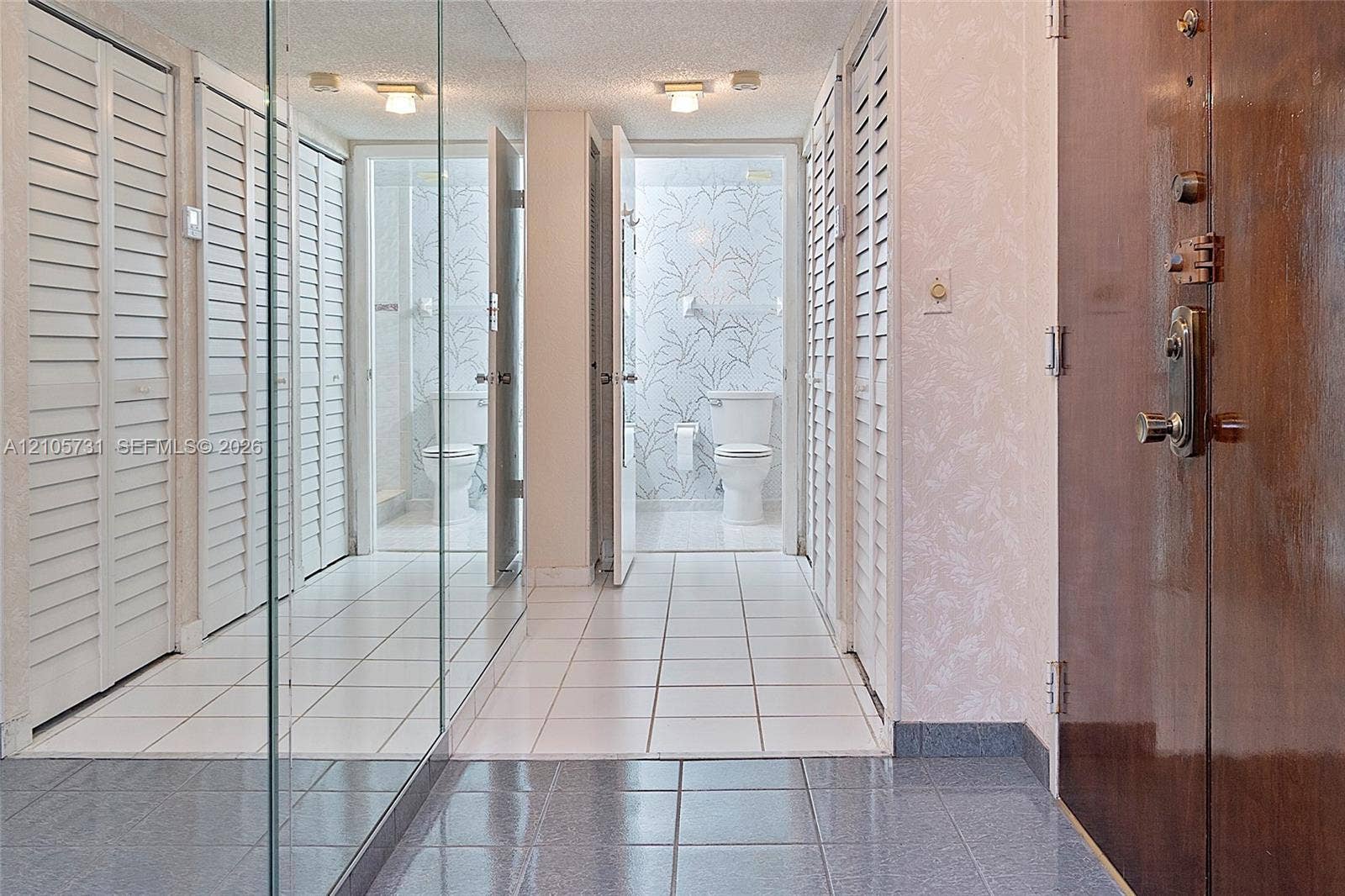5700 SW 127th Avenue, #1404, Miami, FL 33183 Hallway featuring floor-to-ceiling mirror paneling, white louvered closet doors, square tile flooring, and flush-mount ceiling lighting