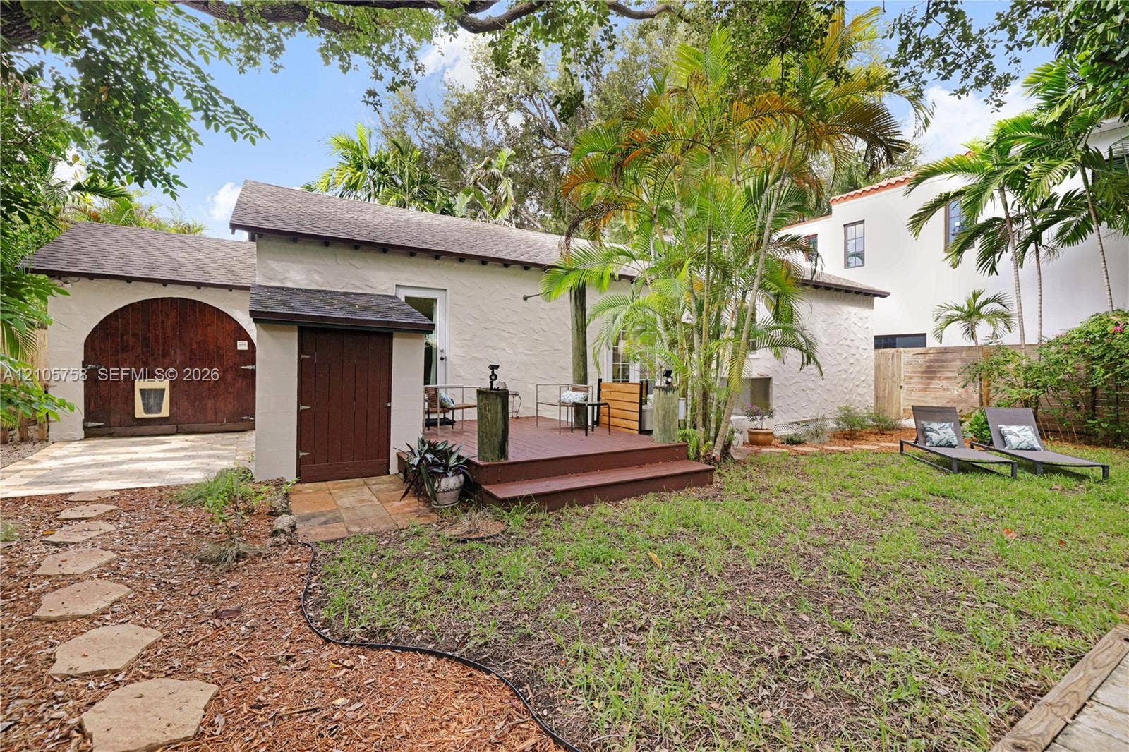 668 NE 70th Street, Miami, FL 33138 Stucco exterior featuring an arched wooden carriage door, raised wood-finish deck, stone paver pathway, and mature landscaping
