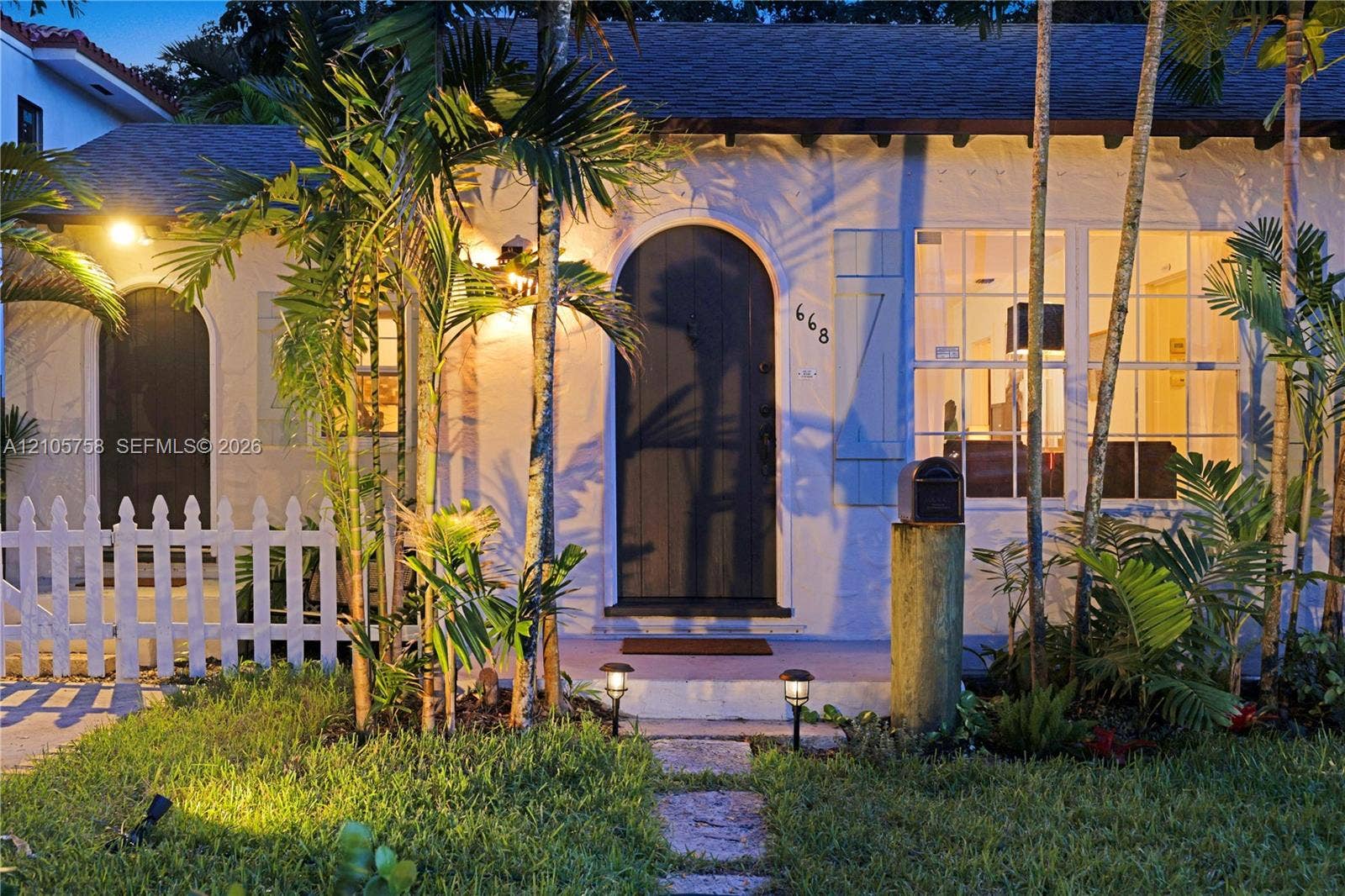 668 NE 70th Street, Miami, FL 33138 Stucco exterior featuring an arched entryway, shuttered multi-pane windows, a stone walkway, and a white picket fence