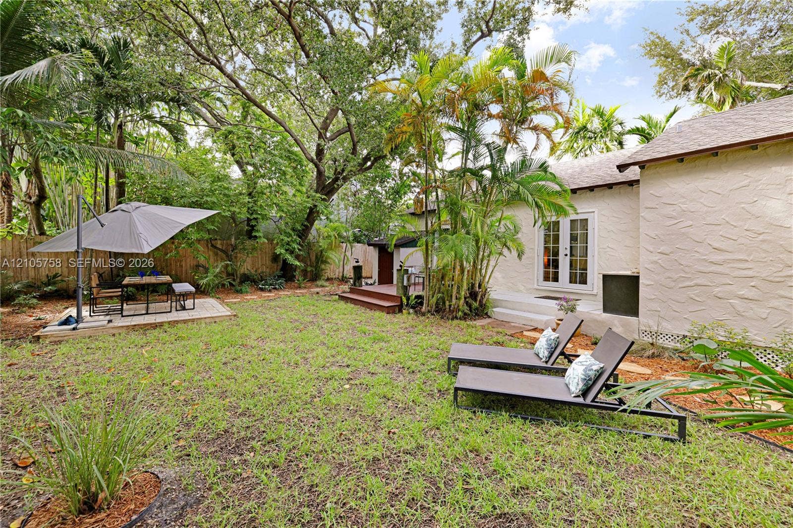 668 NE 70th Street, Miami, FL 33138 Rear exterior featuring a textured stucco facade, double glass-paneled doors, a raised wooden deck, a paved stone patio, and mature landscaping