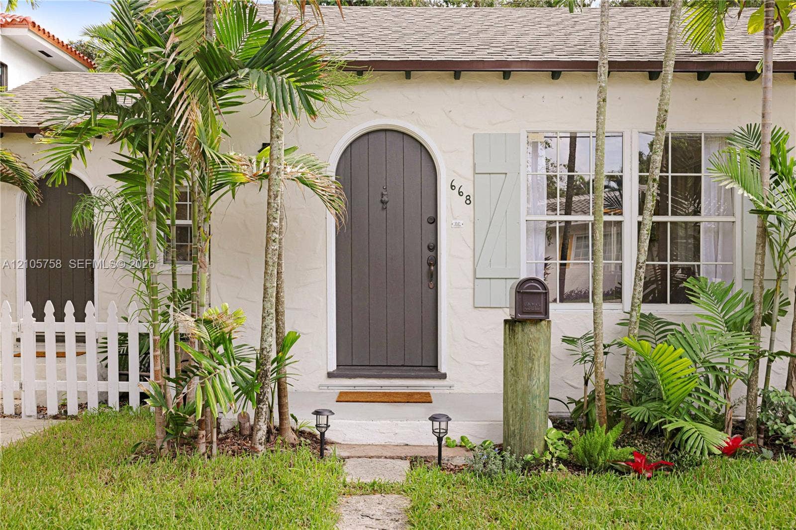 668 NE 70th Street, Miami, FL 33138 Exterior facade featuring stucco siding, an arched entry door, paneled shutters, and a white picket fence