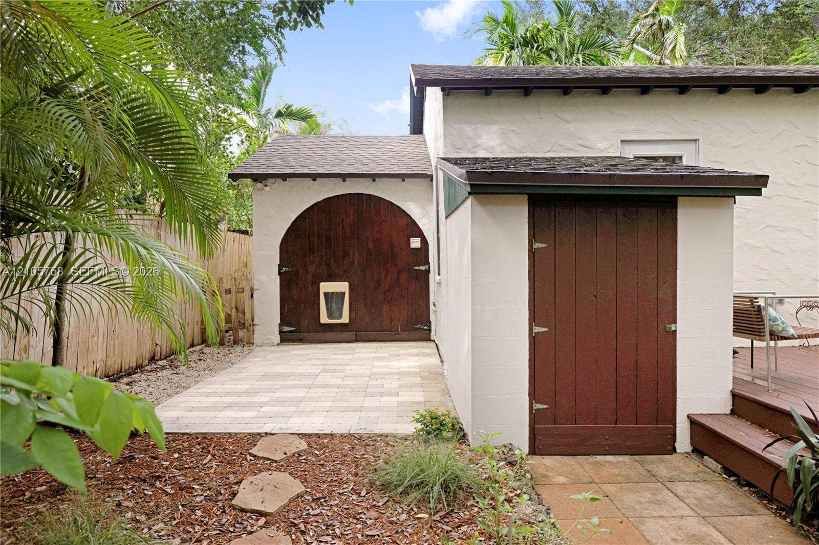 668 NE 70th Street, Miami, FL 33138 Stucco exterior featuring a distinctive arched wood door, a separate wood-panel utility entry, paved stone patio, and wood decking