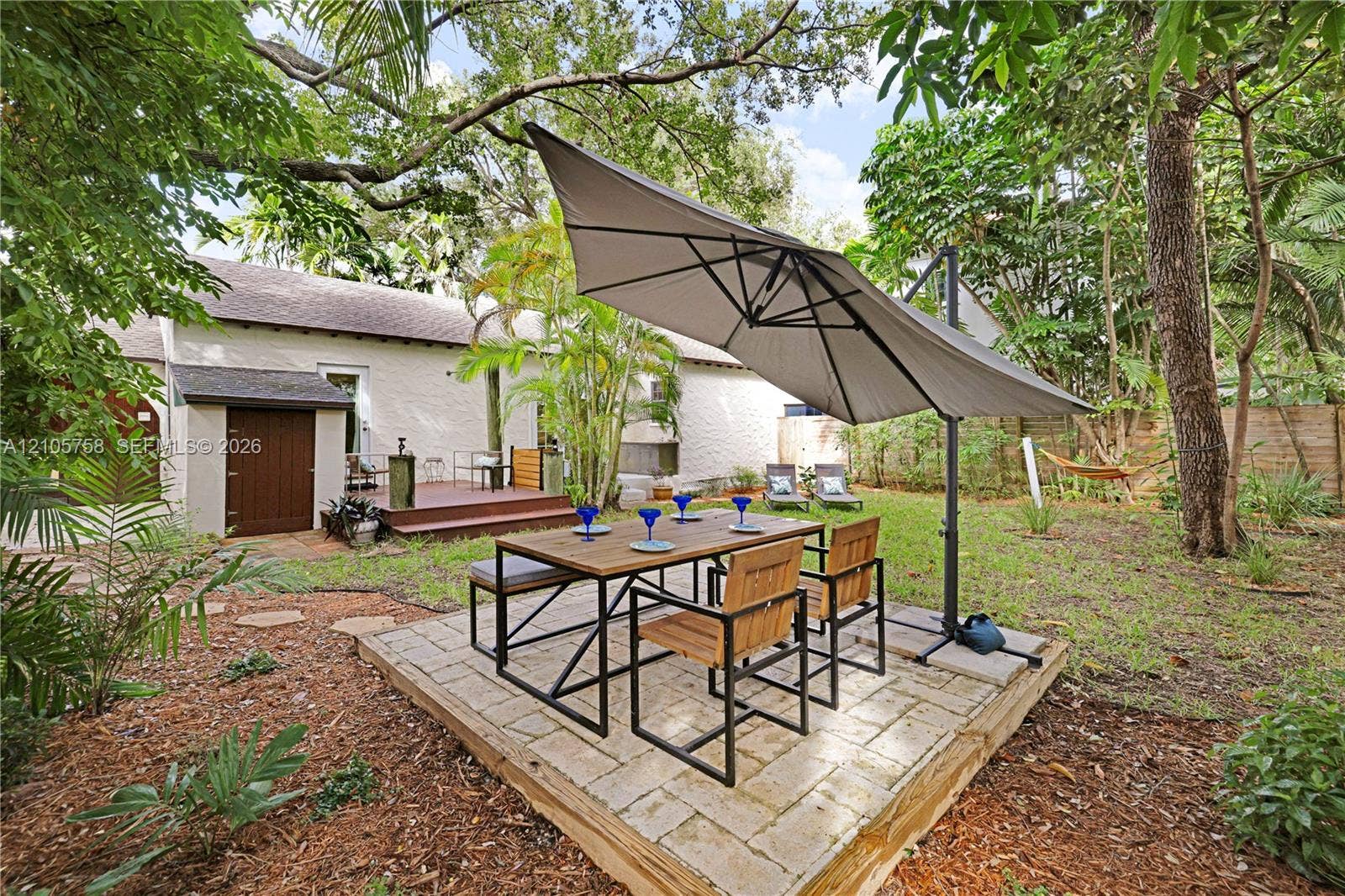 668 NE 70th Street, Miami, FL 33138 Backyard featuring a raised stone paver patio, elevated wood-decking platform, mature shade trees, and wood-finish exterior door